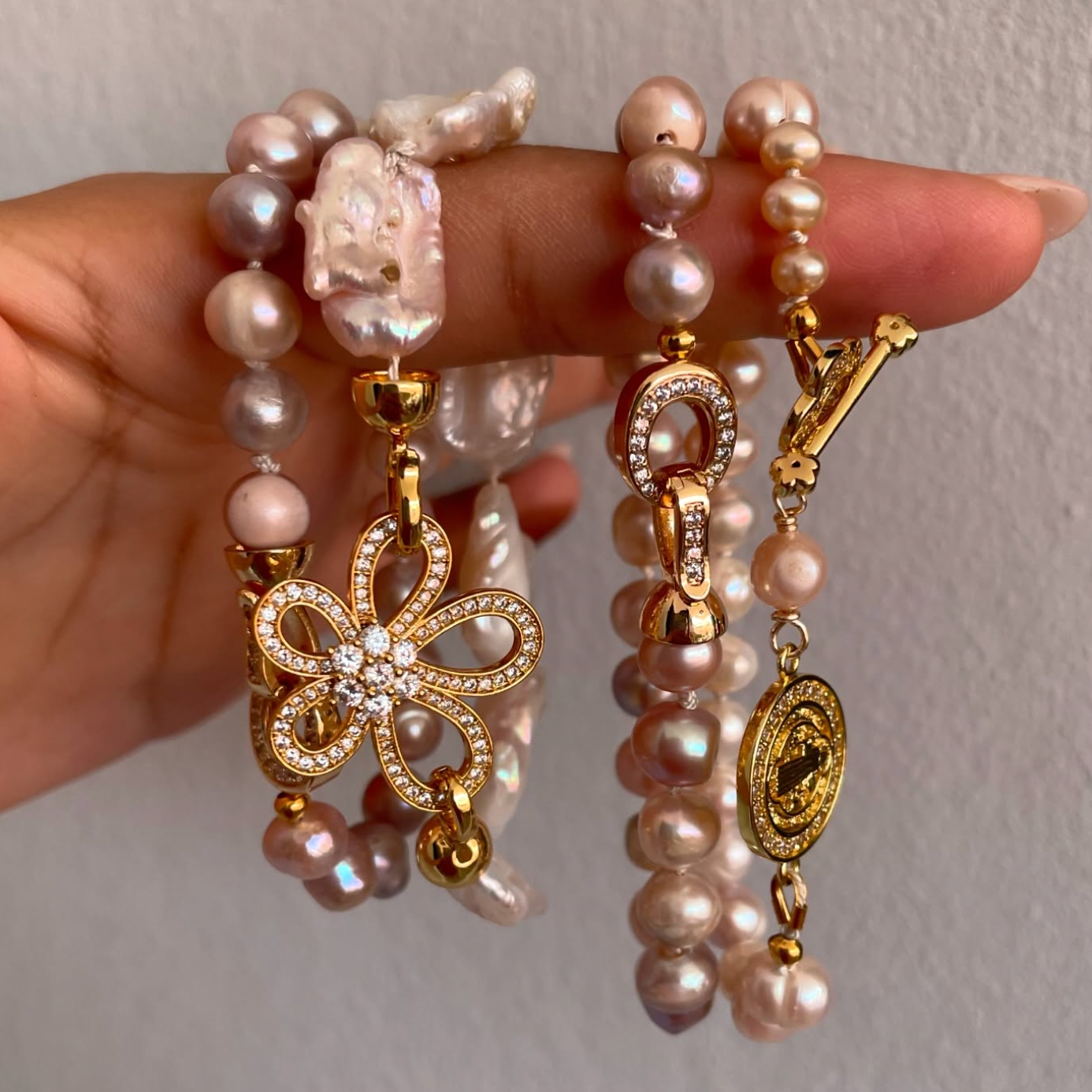 Hand holding multiple pearl and gold necklaces with decorative pendants, including a butterfly, a horseshoe, and an oval medallion.