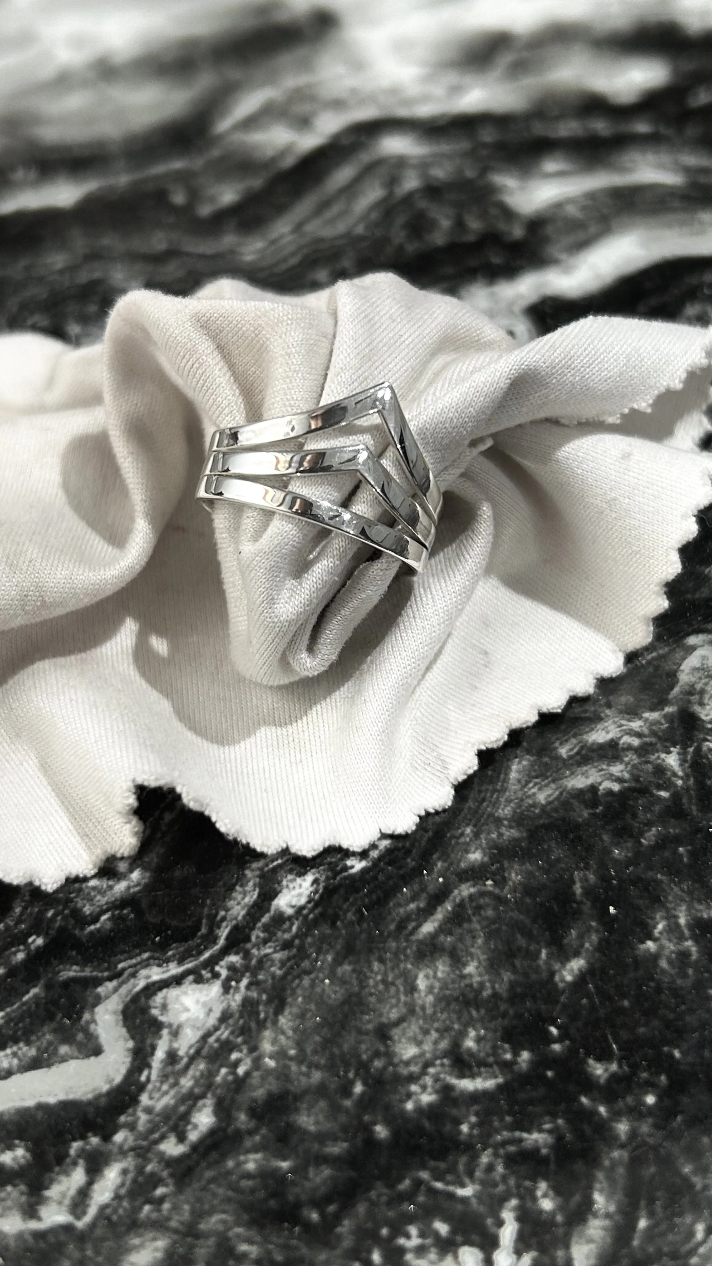 A silver ring with geometric design, resting on a white cloth, on a dark, marbled surface.
