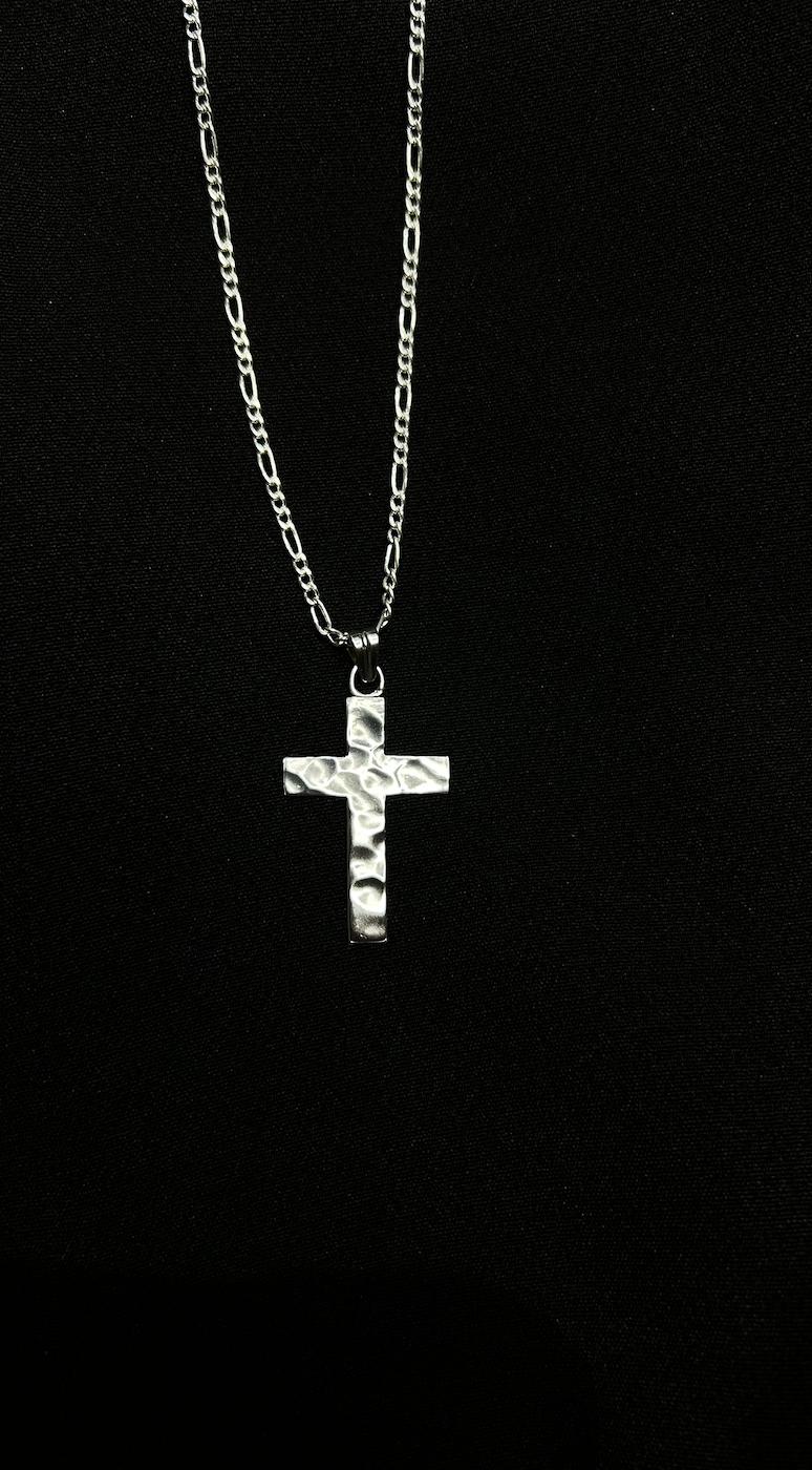 Silver cross necklace with textured surface hanging from a chain against a black fabric background.