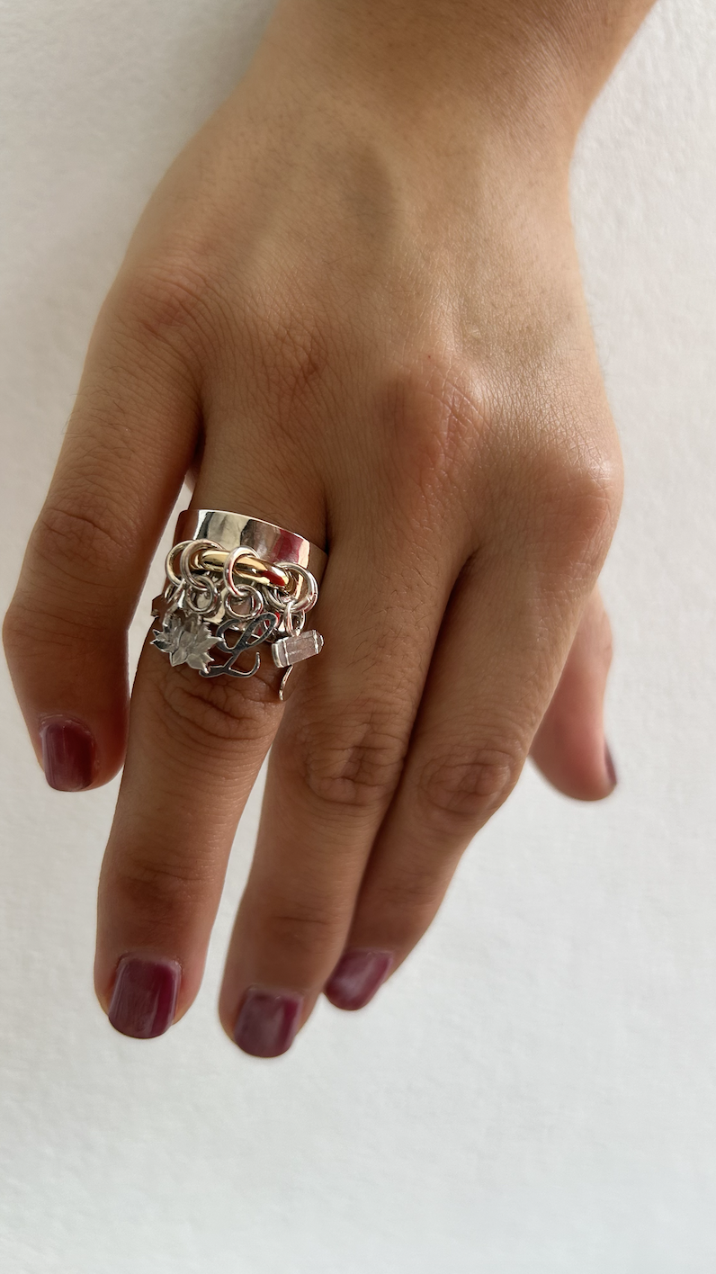 Close-up of a hand with a silver ring that has multiple charms and embellishments, against a plain light background.