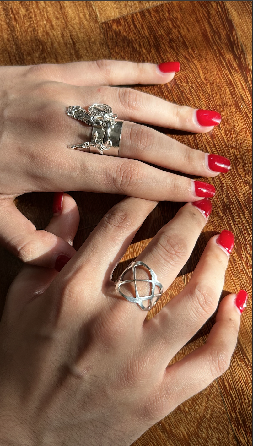 Two hands with red-painted nails, each wearing rings, resting on a wooden surface. One hand has a silver ring with a pentagram design, and the other has a silver ring with a chain-like design.
