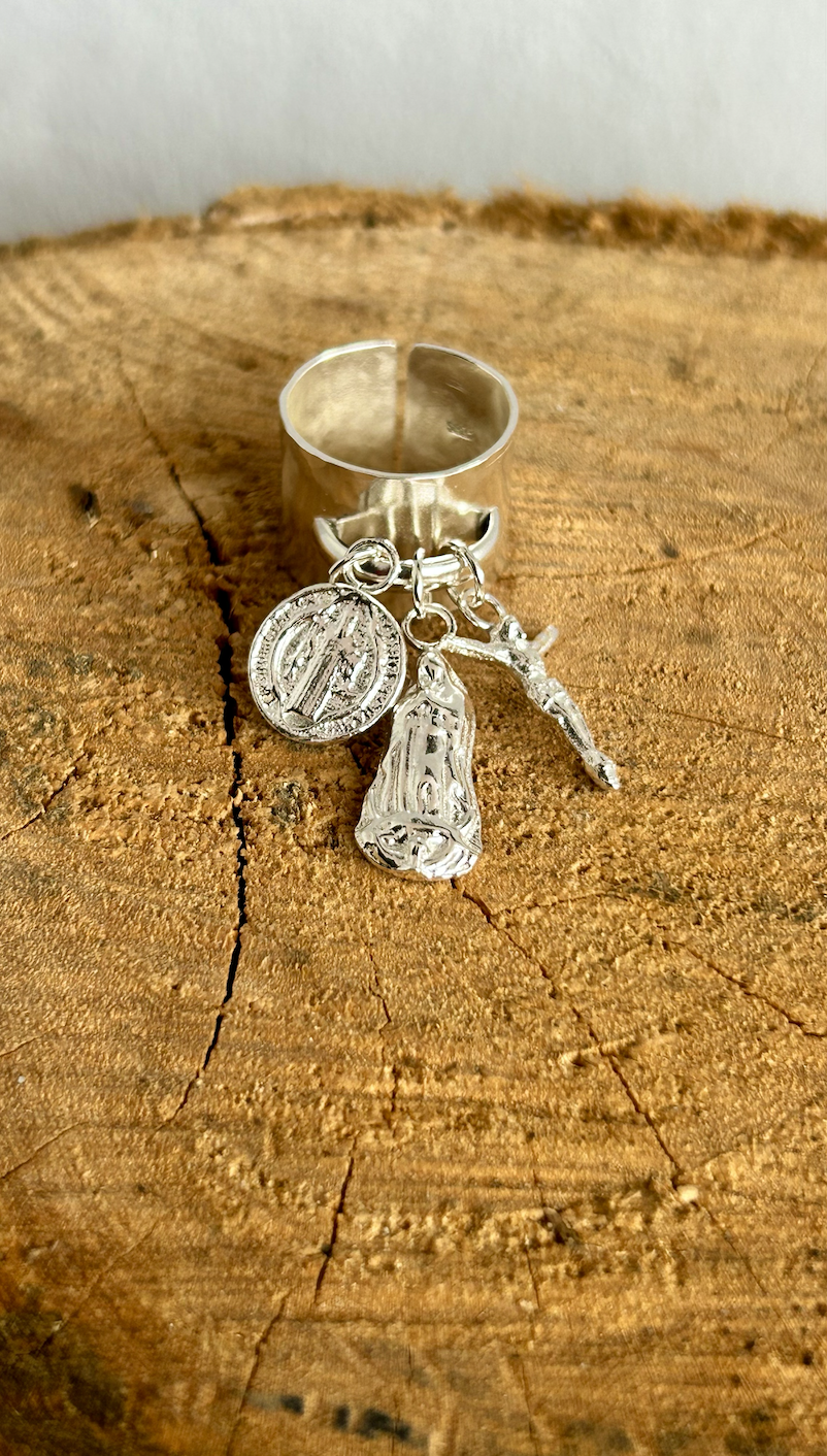 A silver ring with three attached charms on a wooden surface. The charms include a medallion, a figure, and a small sword.