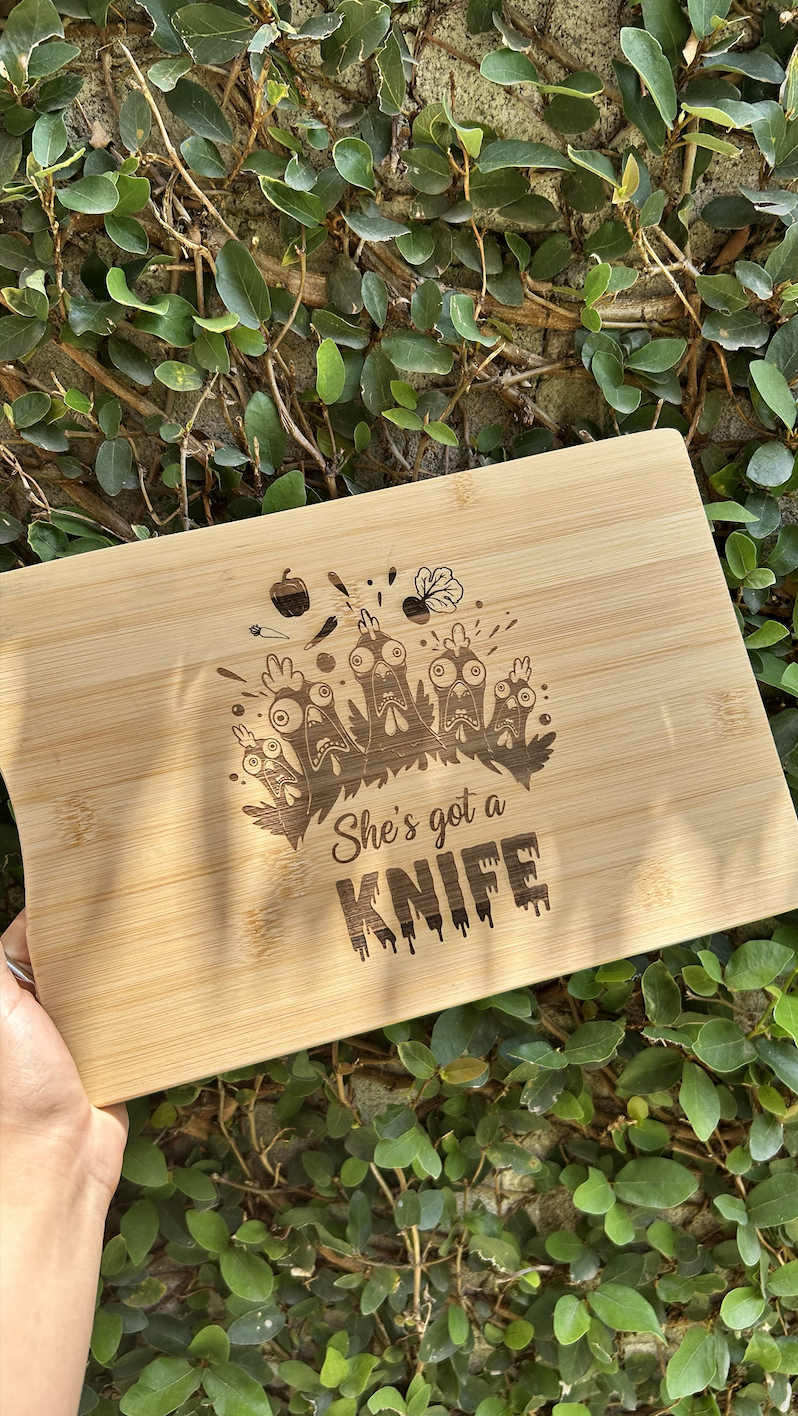 A wooden cutting board with a humorous design featuring angry cartoon chickens and the phrase "She’s got a KNIFE," held above a background of green leafy plants.