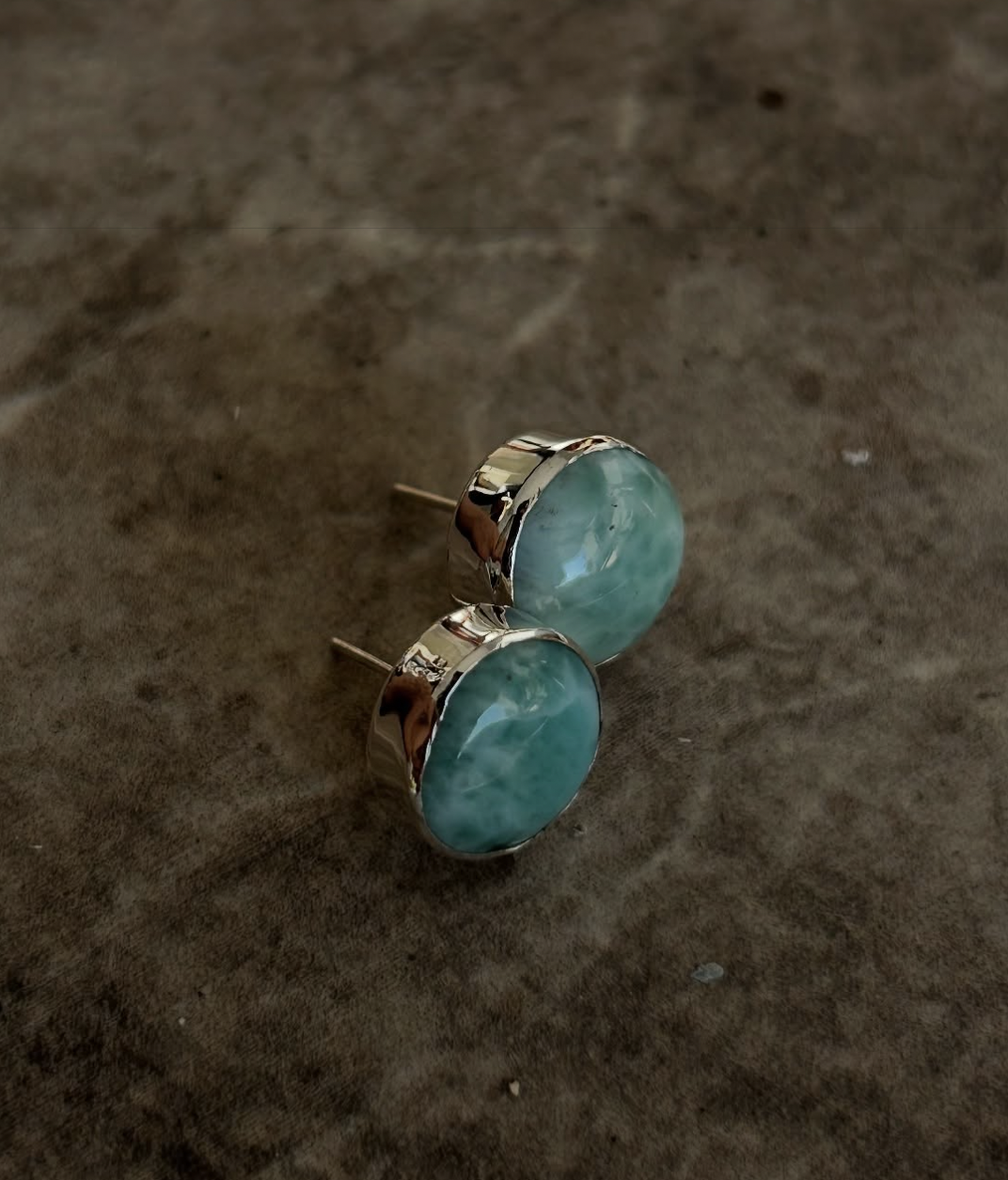 A pair of round, turquoise-colored earrings with silver settings on a dark surface.