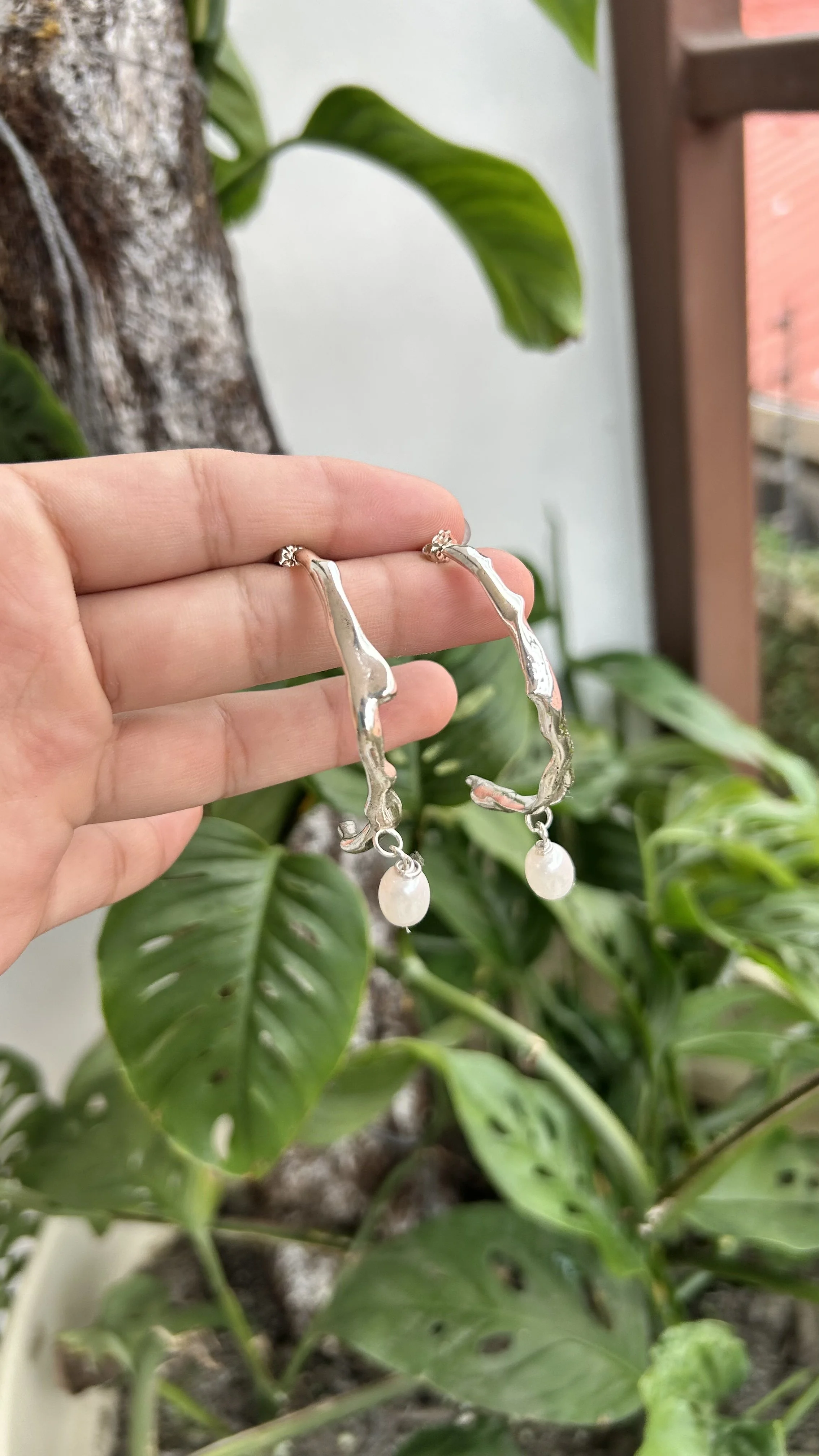 Silver earrings with pearl drops held against a background of green leaves.
