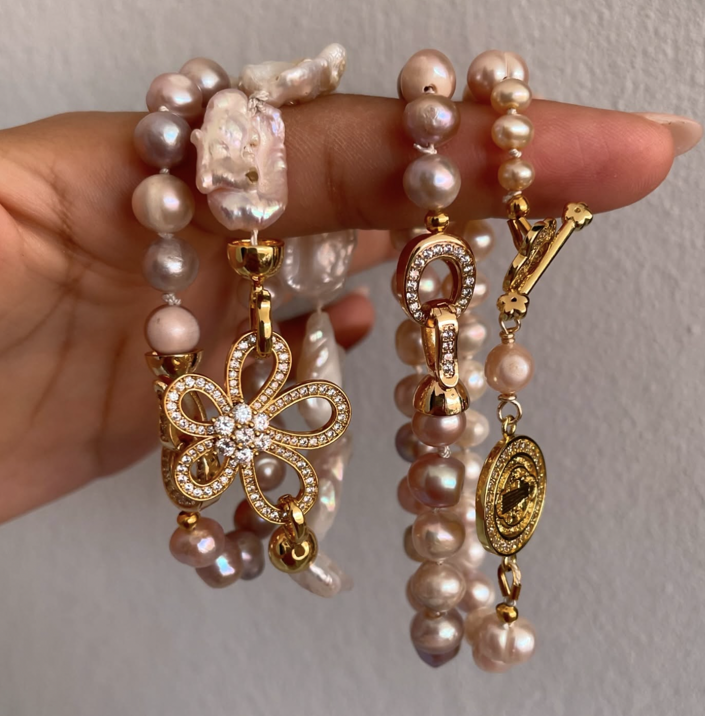 Hand holding multiple pearl necklaces with gold and rhinestone charms, including a butterfly-shaped charm, a horseshoe-shaped charm, and an oval-shaped charm against a plain light background.