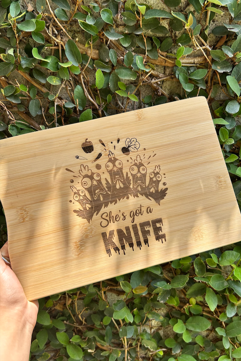 A wooden cutting board with a humorous design that says "She's got a KNIFE" accompanied by cartoonish angry chickens and various cooking-related icons, held against a background of green leaves and a brick wall.