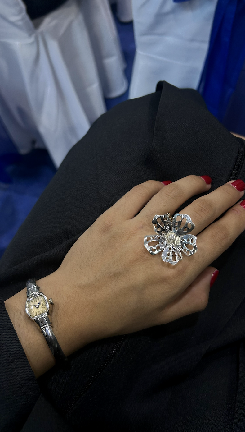A person's hand with red painted nails resting on their lap, wearing a large silver butterfly-shaped ring and a silver wristwatch, with a background of white and blue fabric.
