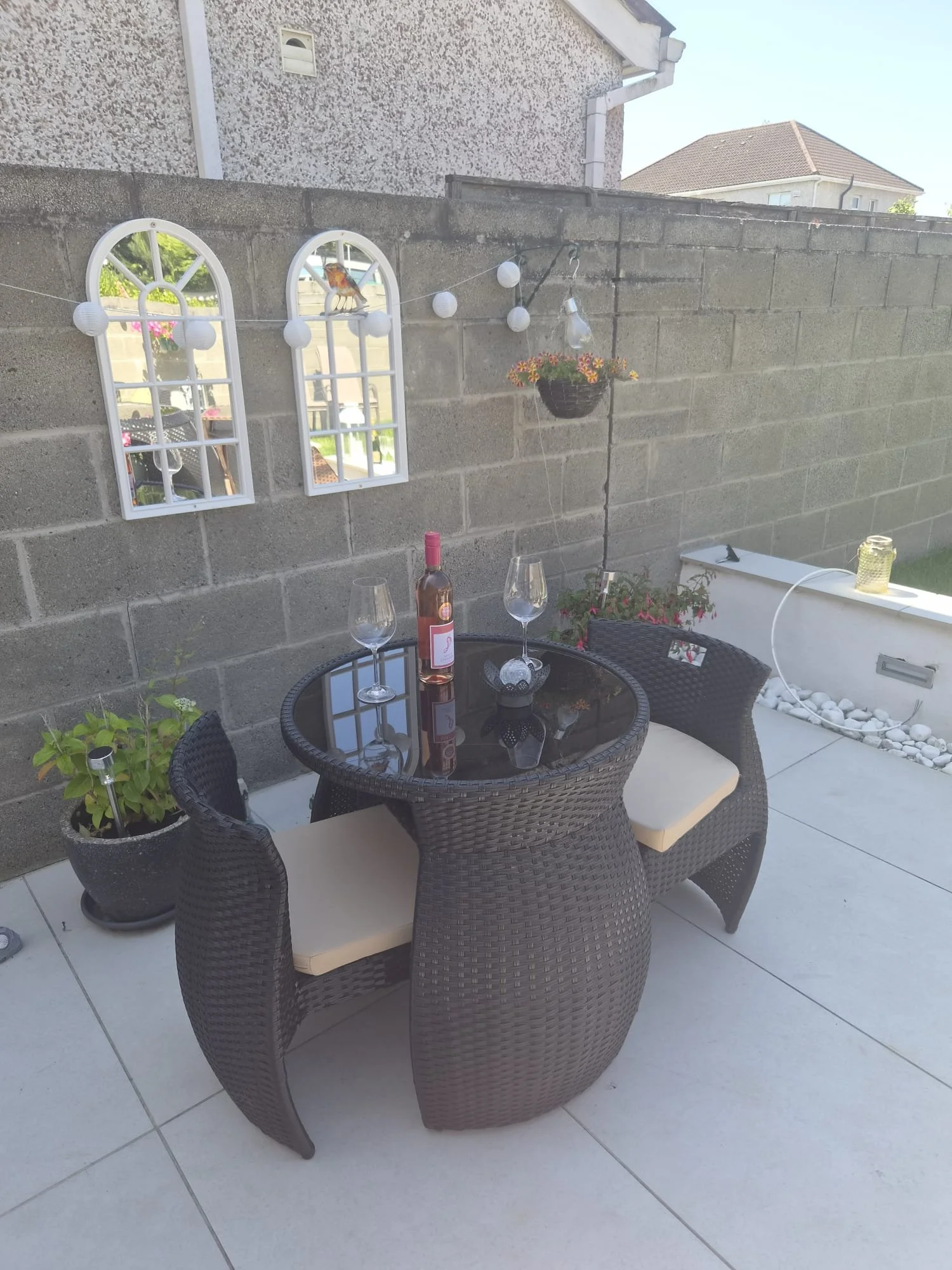 An outdoor patio area with a round glass-top table surrounded by five wicker chairs with cushions. There is a bottle of rosé wine and two empty glasses on the table. The background features a concrete block wall decorated with white window-shaped frames, hanging string lights, a hanging flower basket, and other potted plants. The patio has tiled flooring, and neighboring houses with similar tiled roofs are visible.