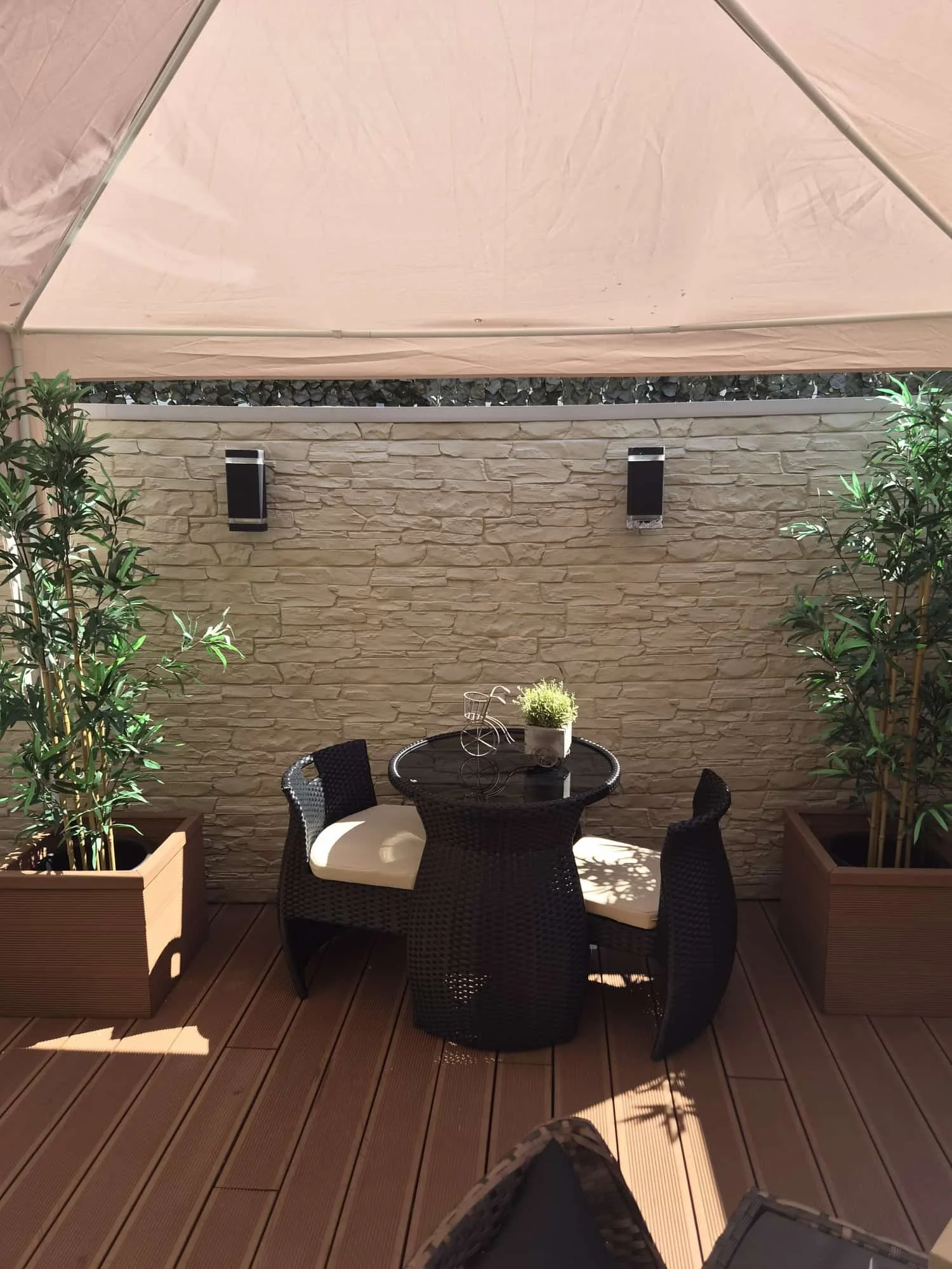 View of an outdoor patio with a round glass table, four black wicker chairs with cushions, potted plants, and a beige brick wall under a canopy.
