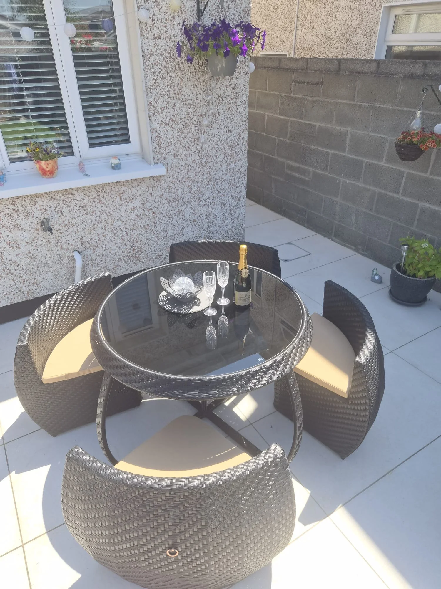 Outdoor patio table with champagne bottle and two glasses surrounded by four wicker chairs, with a textured house wall, decorated with potted plants and hanging flower basket.