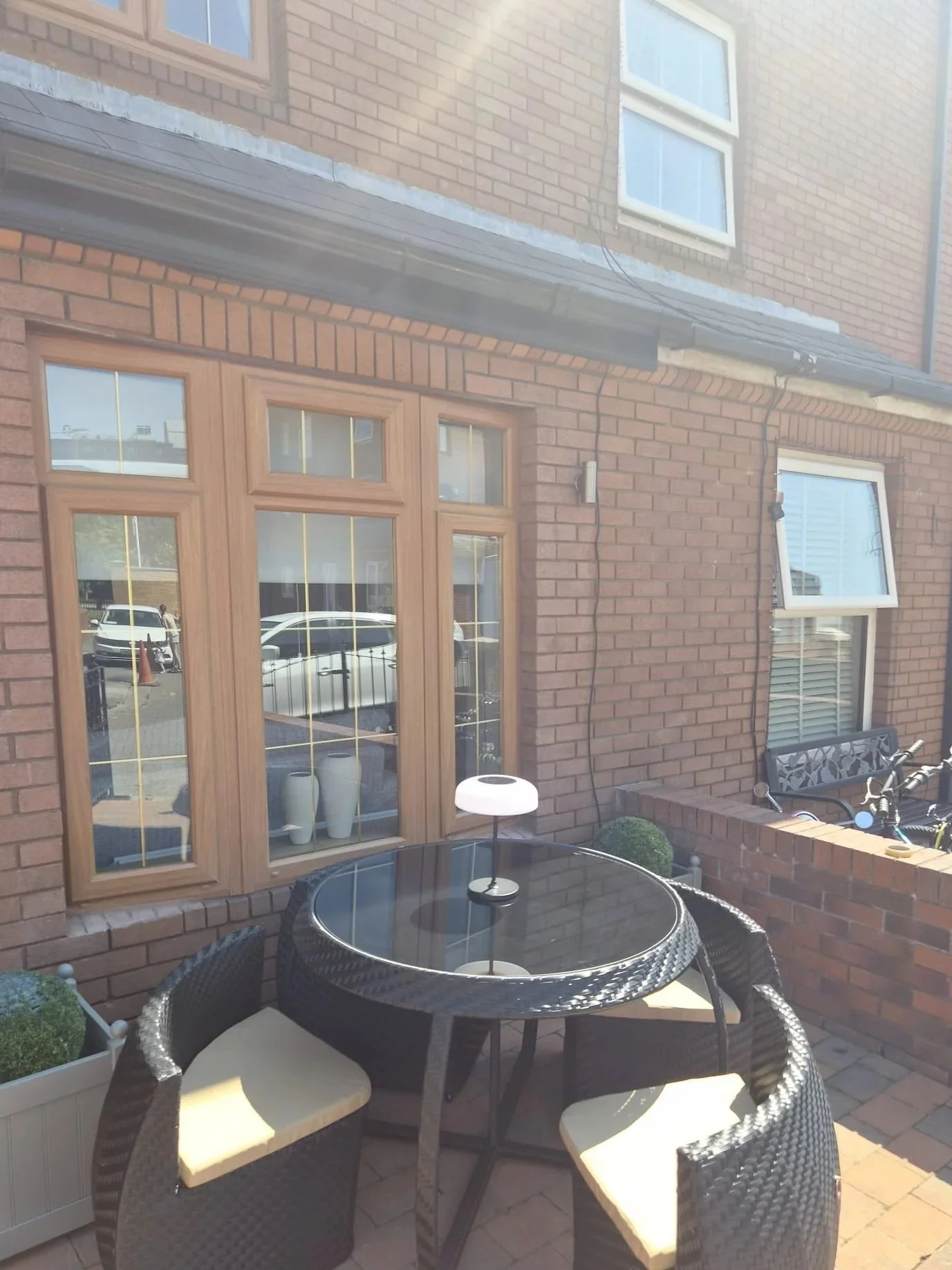 A small outdoor patio with a round glass table surrounded by four wicker chairs with beige cushions. There are two white planters with green plants, a black metal bench, and an orange brick house in the background with multiple windows and a door.