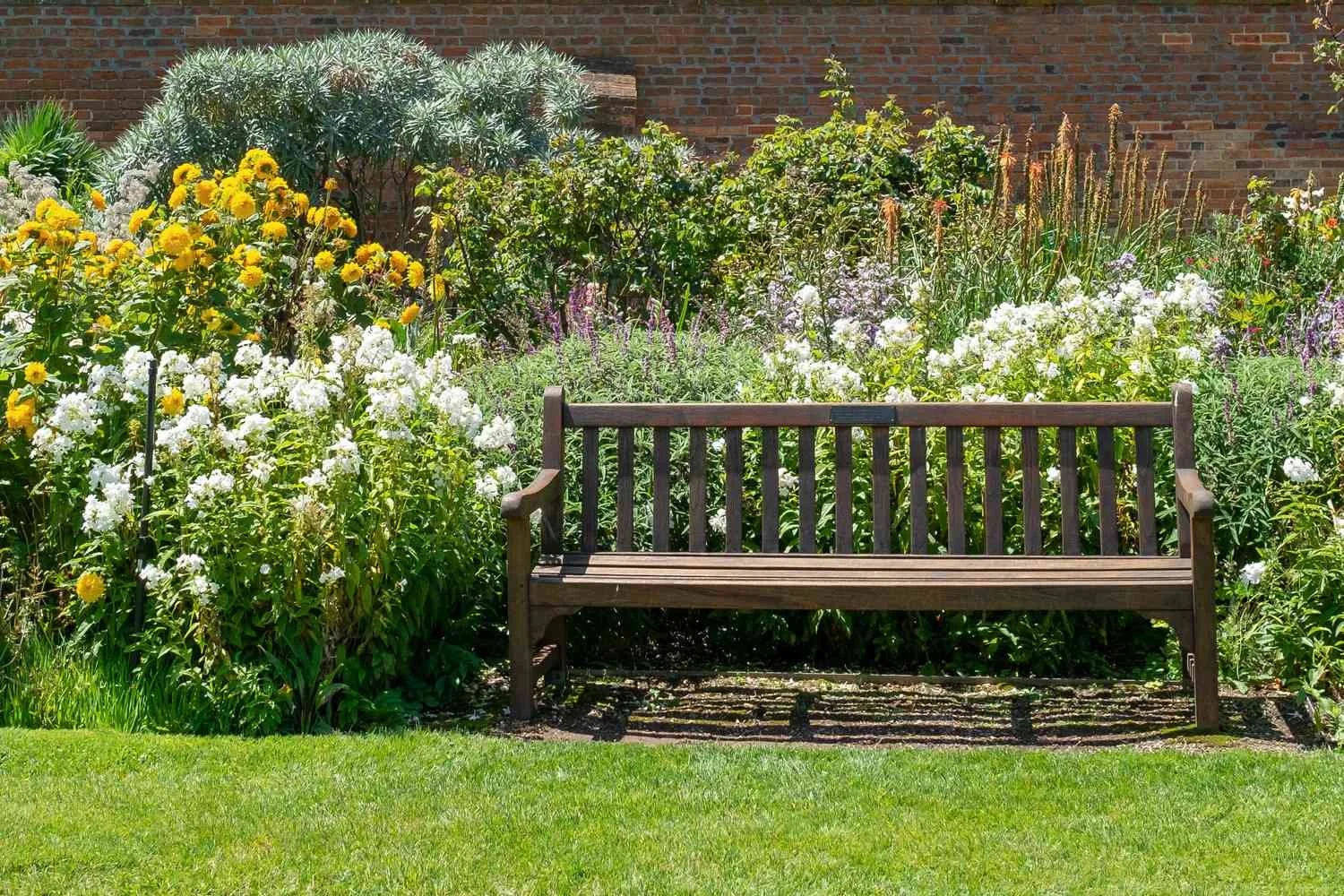 A wooden bench in a garden surrounded by colorful flowers and greenery, with a brick wall in the background.