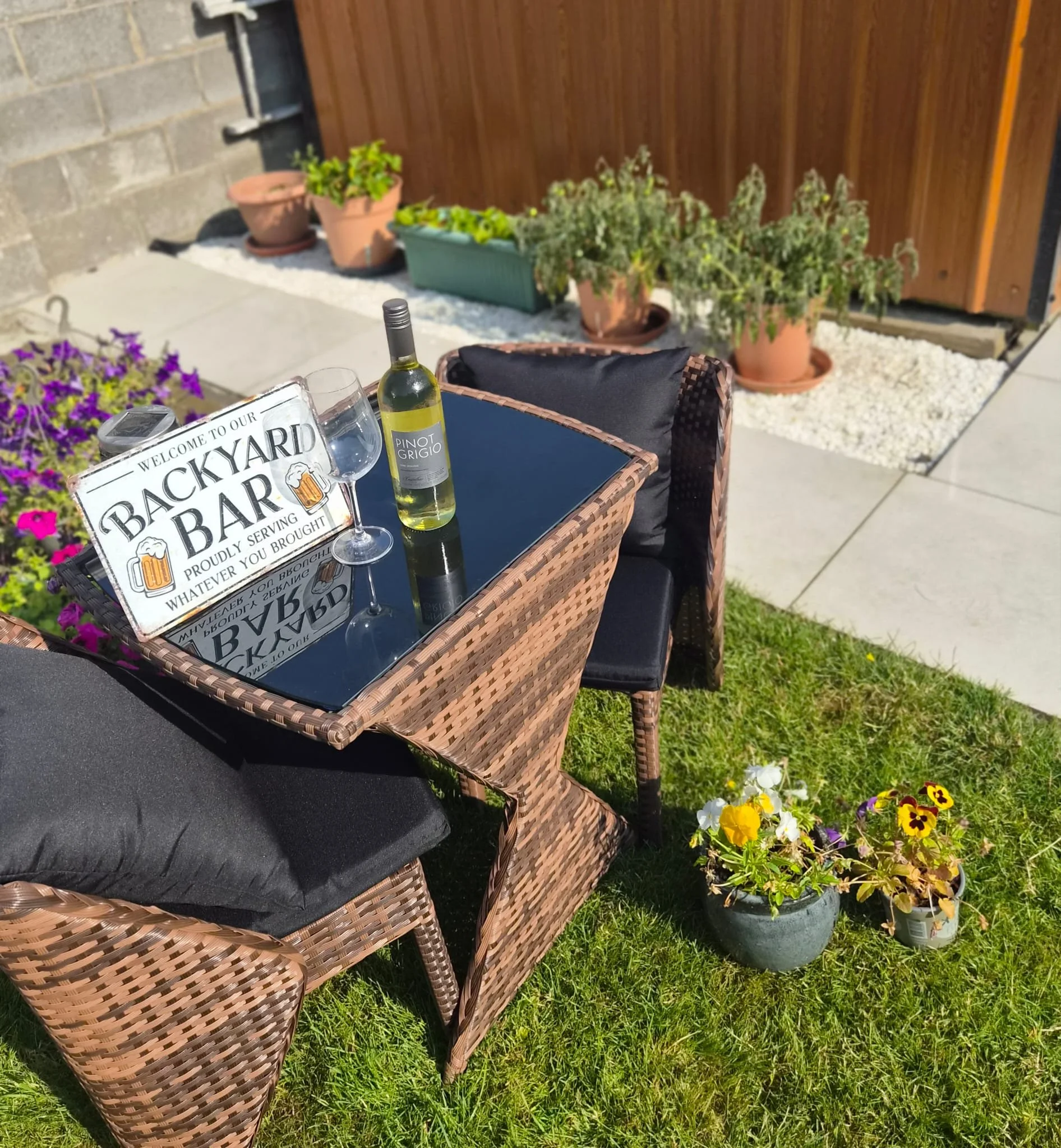 Outdoor patio scene with a wicker table set for a gathering, featuring a sign that says 'Backyard Bar' and a bottle of Pinot Grigio wine. There are wine glasses, and potted plants with colorful flowers surround the area. The background has a stone wall and a wooden fence with additional potted plants.