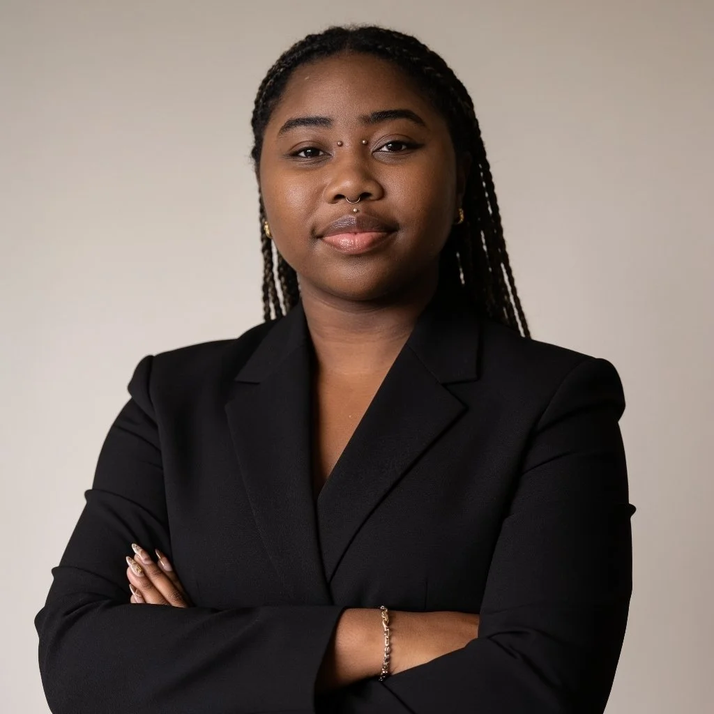 A confident African American woman with braided hair and nose piercings, wearing a black blazer, standing with arms crossed against a neutral background.