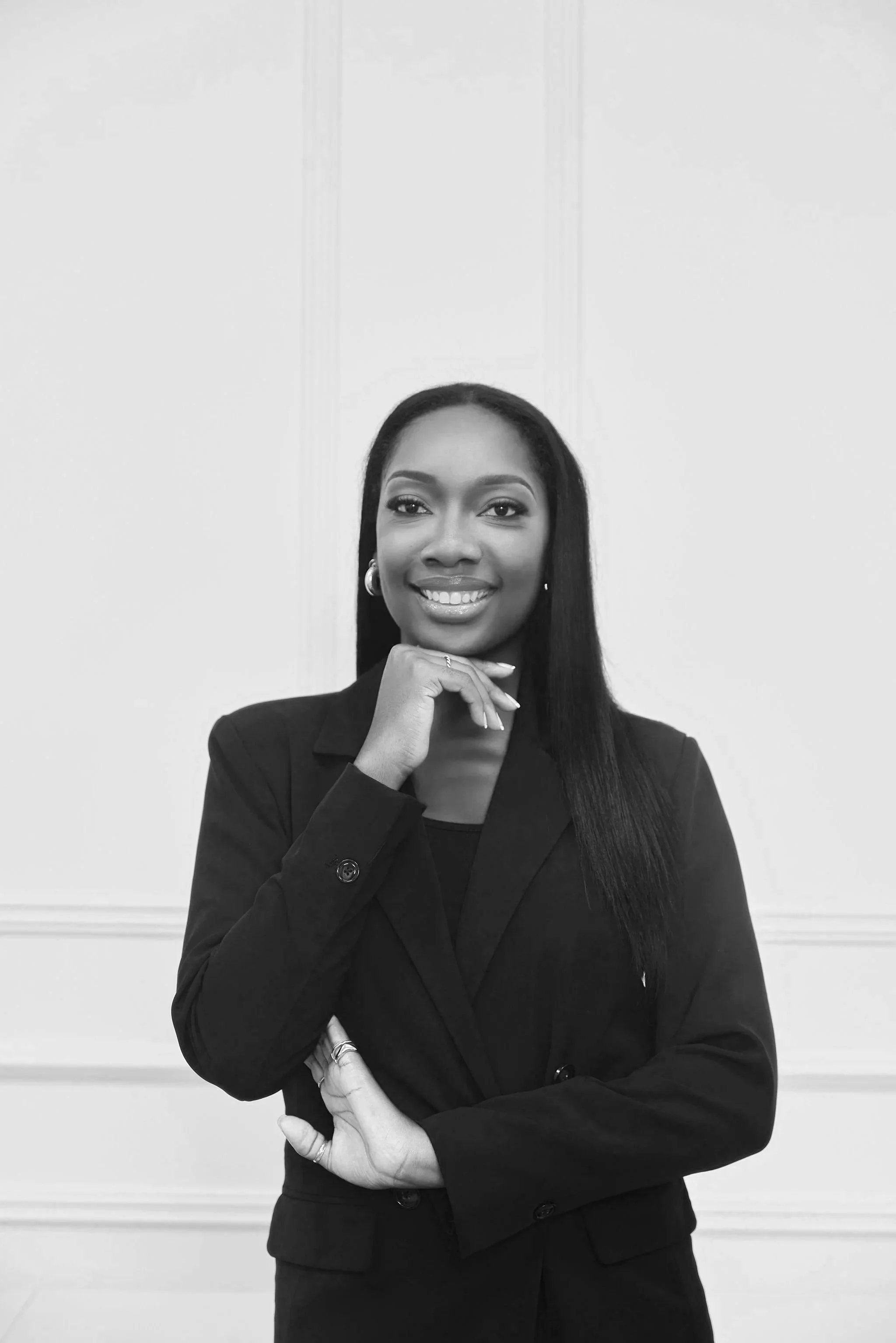 A confident female lawyer with long, straight hair and a bright smile, wearing a dark blazer, standing against a plain wall with a decorative mold in the background.