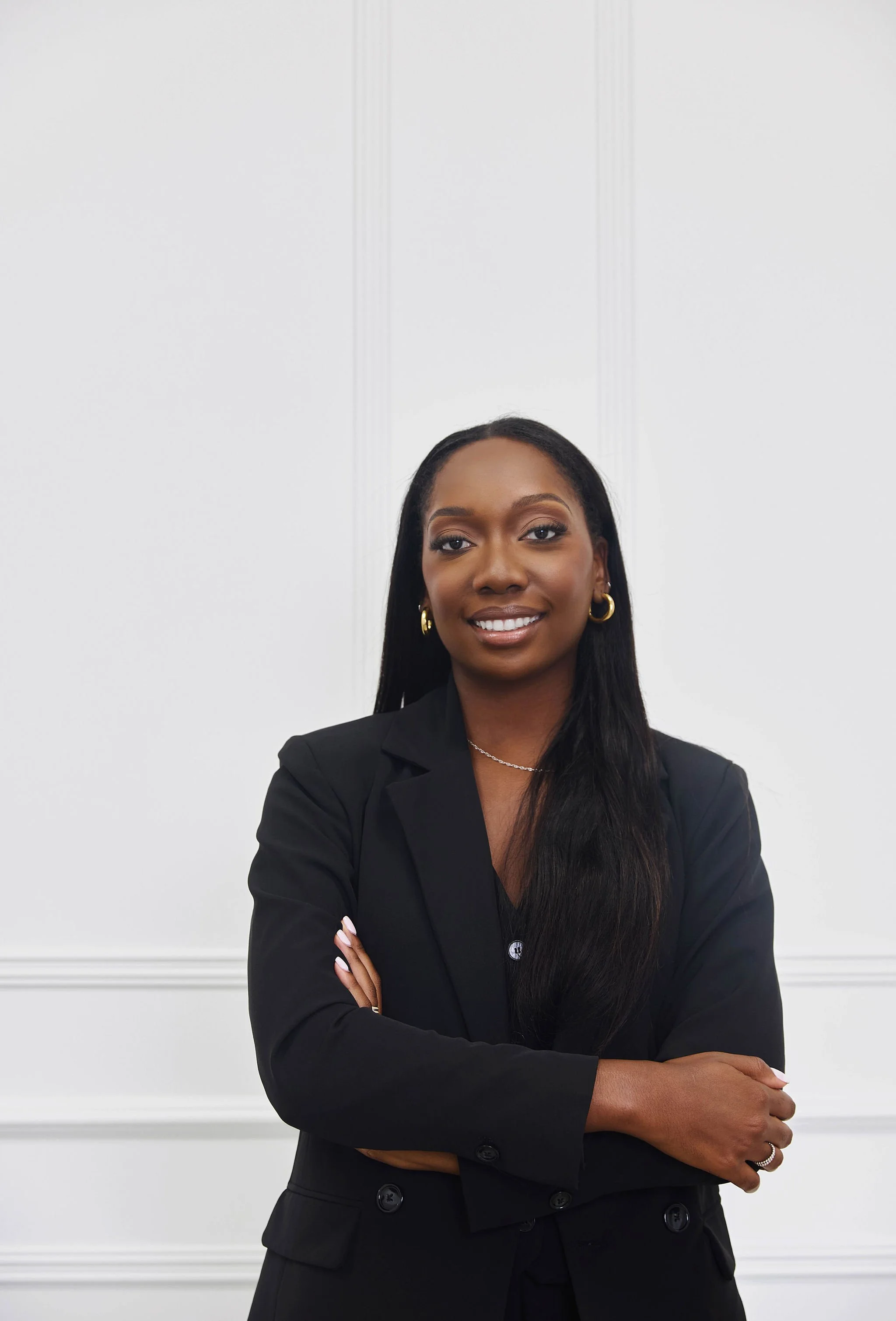 A confident Black female lawyer with long black hair and gold hoop earrings, wearing a black blazer, standing with her arms crossed in front of a white wall with decorative moldings.