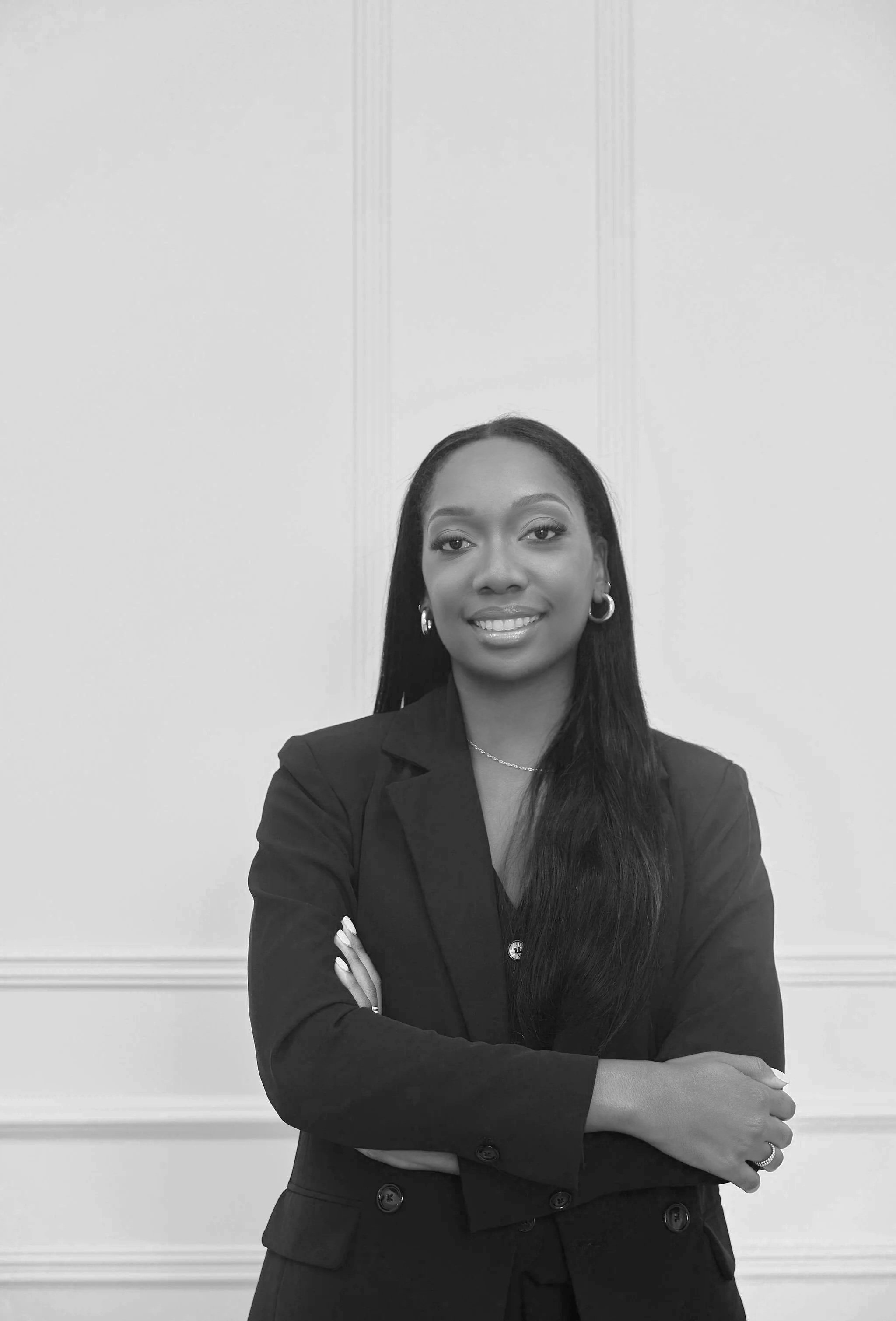 A confident Black female lawyer with long black hair and gold hoop earrings, wearing a black blazer, standing with her arms crossed in front of a white wall with decorative moldings.