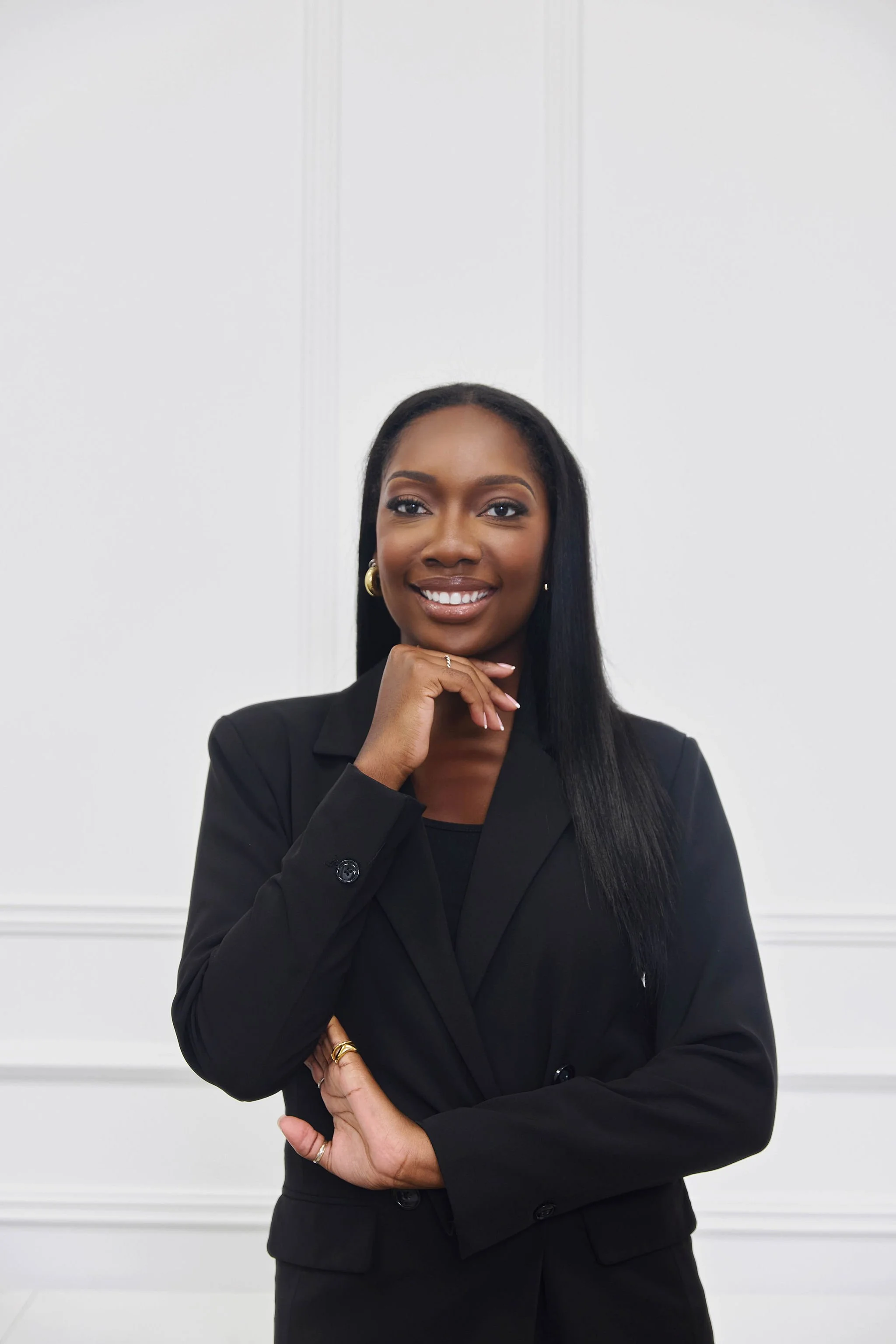 A confident Black female lawyer with long black hair and gold hoop earrings, wearing a black blazer, standing with her arms crossed in front of a white wall with decorative moldings.