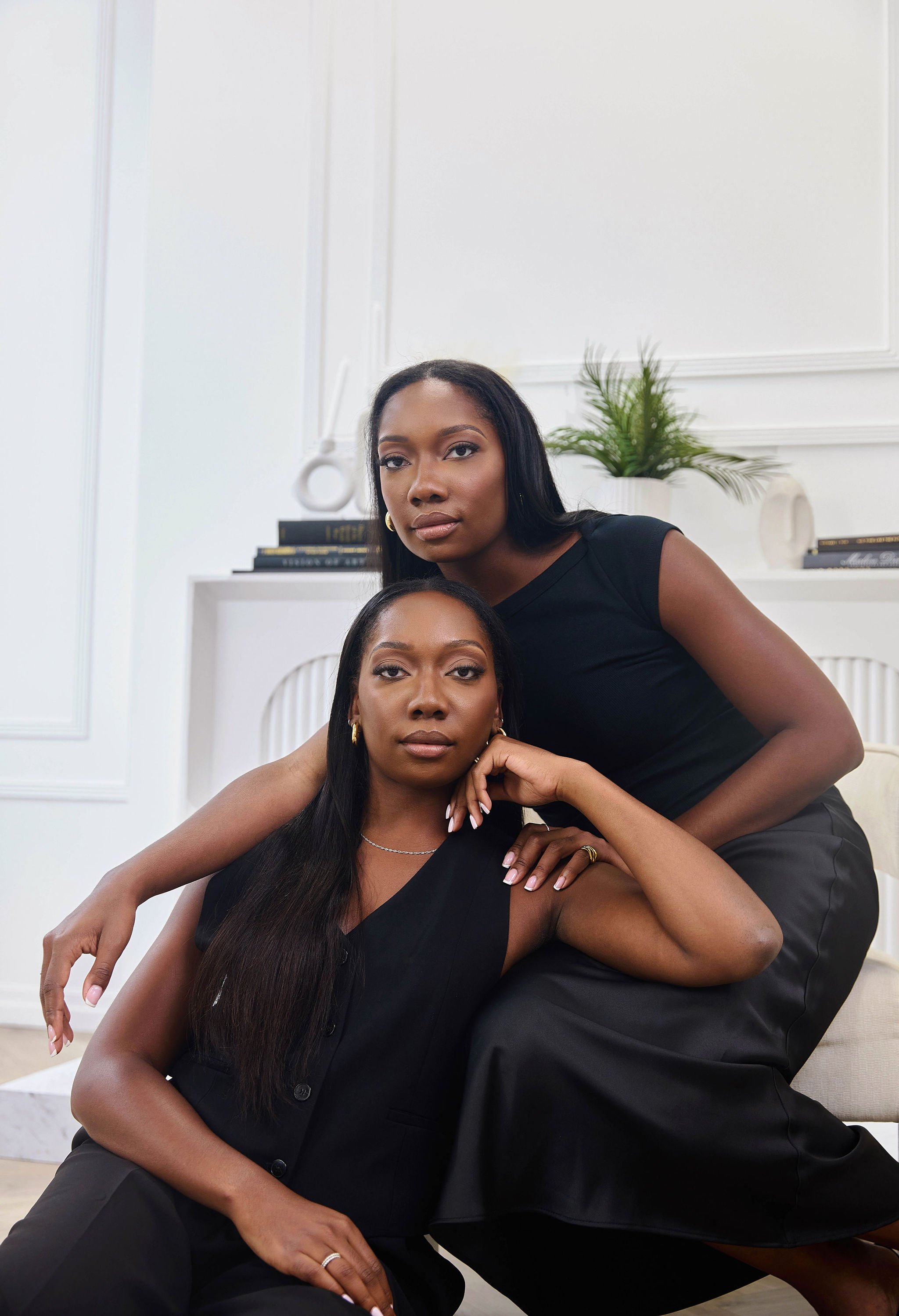 Two female lawyers with dark hair and dark clothing posing indoors, with decorative white walls, green plant, and books in the background.