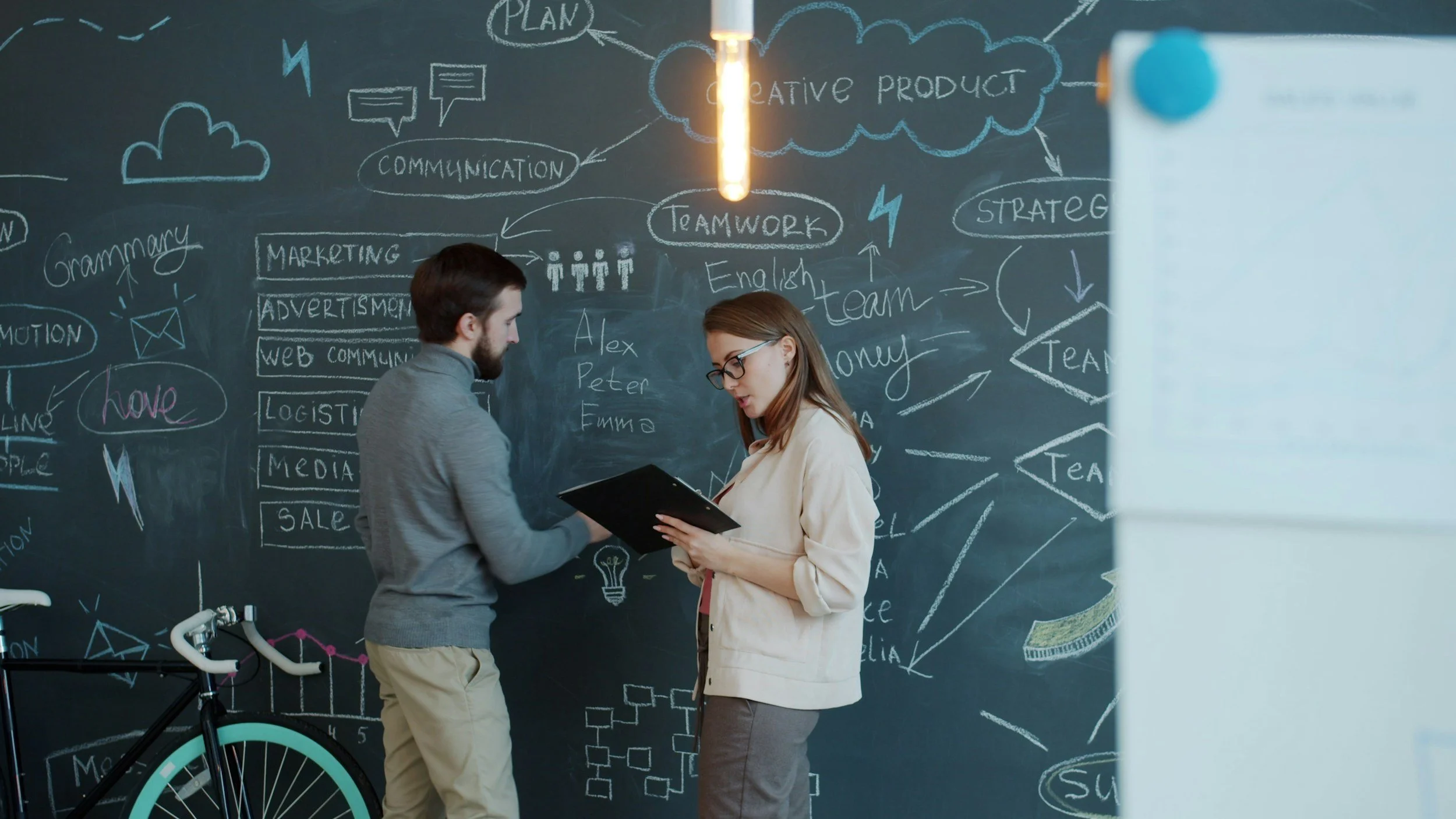 Two people, a man and a woman, are discussing in front of a large chalkboard filled with diagrams and notes related to marketing, teamwork, and creative projects. The woman is holding a clipboard, and there is a bicycle in the foreground.
