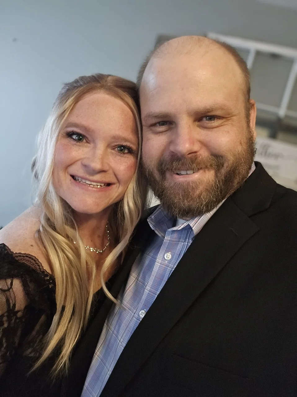 A smiling woman with long blonde hair and a man with a beard in a suit, posing closely together for a photo.