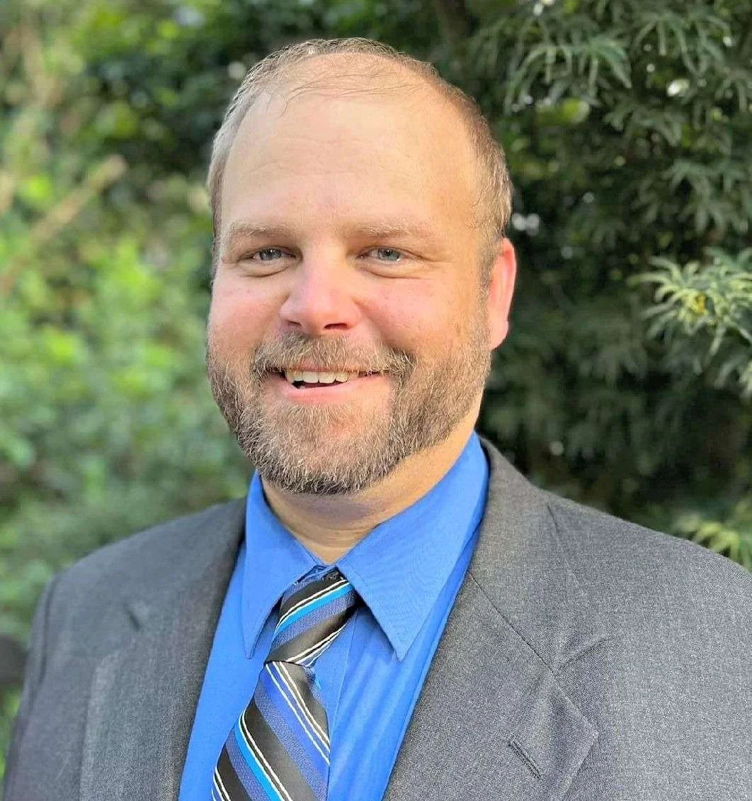 A smiling man in a gray suit, blue shirt, and striped tie standing outdoors with green foliage in the background.
