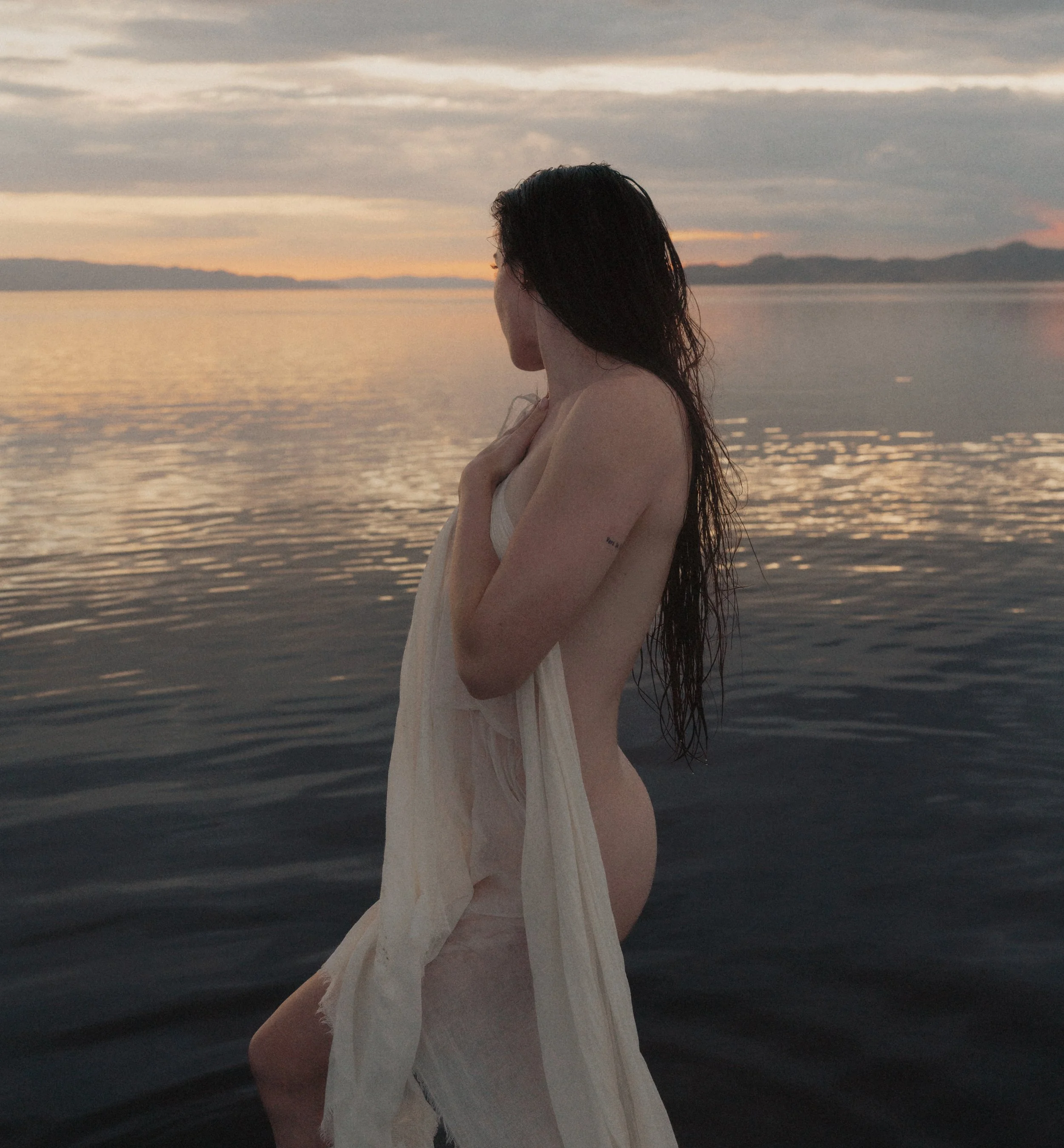 A woman with wet hair and a white dress standing near a body of water during sunset.