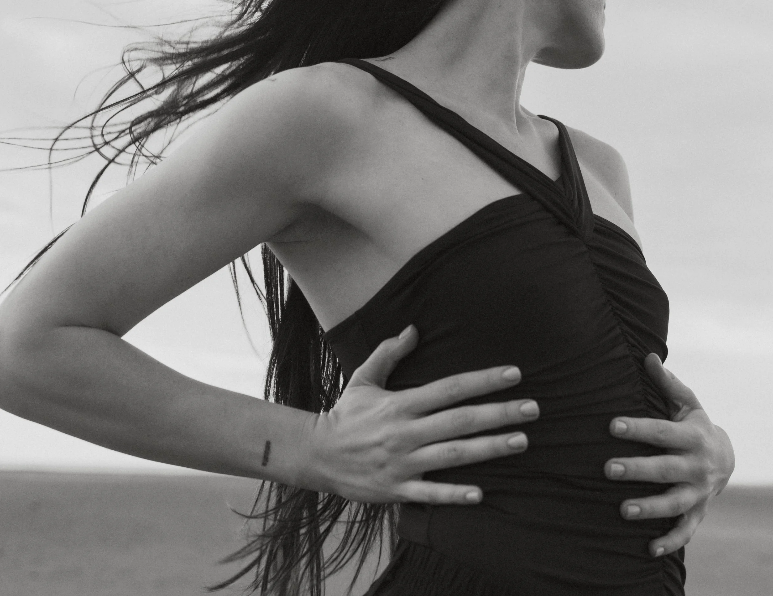 A grayscale image of a woman with long hair, seen from the side, wearing a strappy tank top. Her hand is placed on her waist.