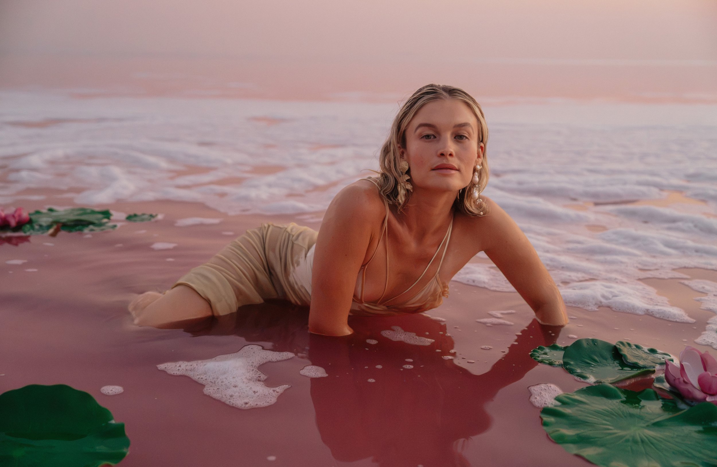 A woman with blonde hair and earrings leaning on her hand in shallow pink water among floating green lily pads and pink flowers, with a pink sky in the background.