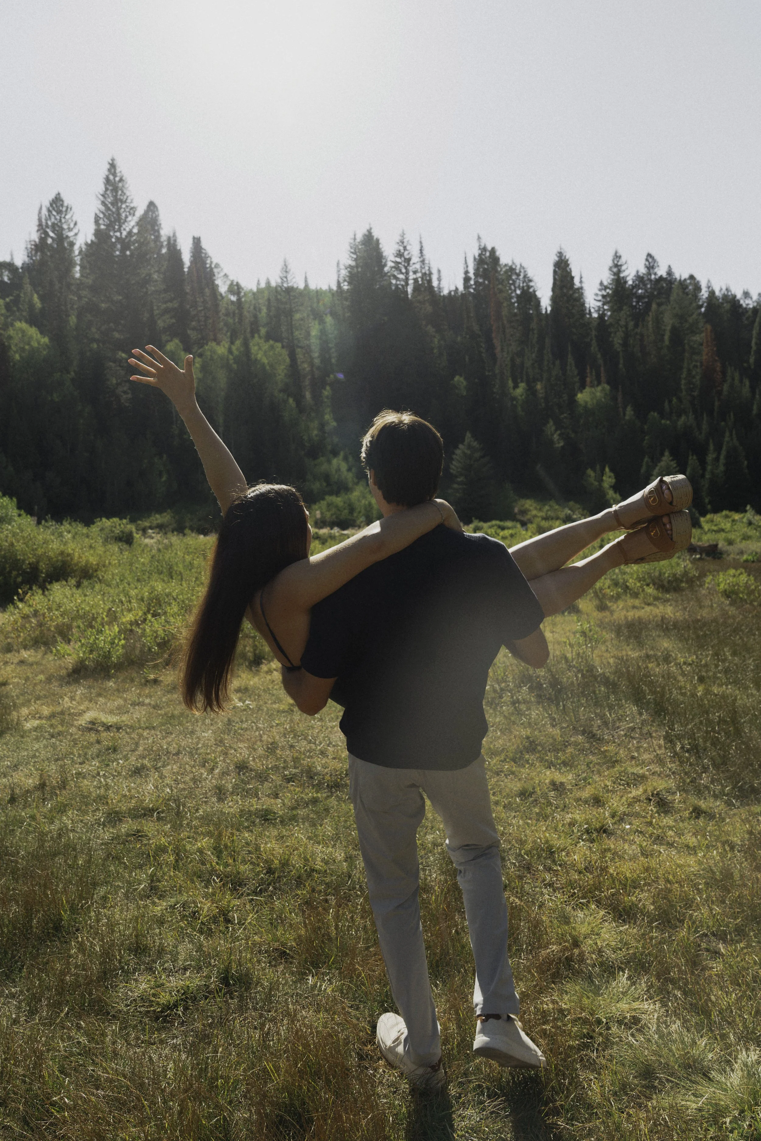 A couple standing in a grassy field with a forest of pine trees in the background. The man is carrying the woman, who has her arm raised and is pointing towards the sky.