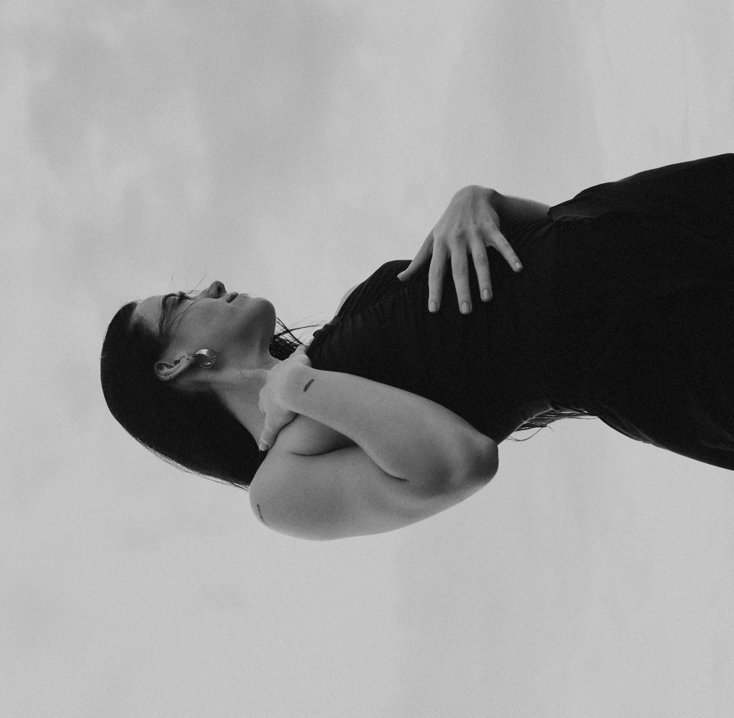 Black and white photograph of a woman lying on her back with one hand resting on her stomach and her eyes closed, with a plain background.