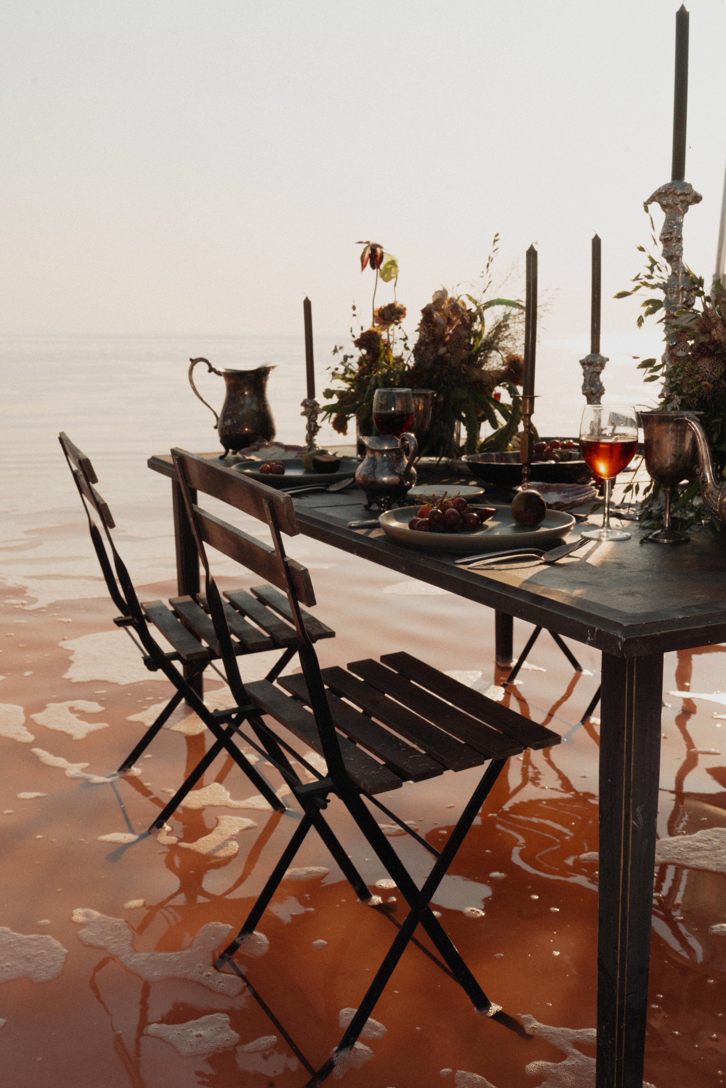 A dining table set near a beach with water touching the shore, adorned with silverware, glasses of red wine, candlesticks, and floral centerpieces, with chairs placed around the table.