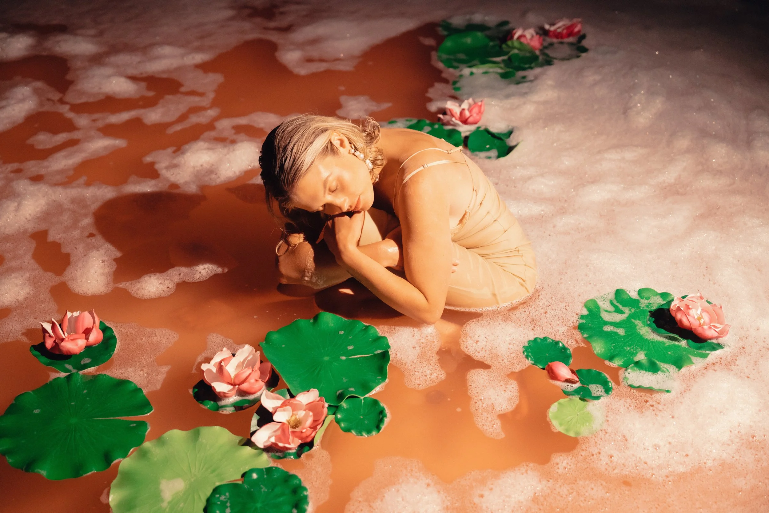 A person with blonde hair and earrings is crouched in pink foam, surrounded by artificial green lily pads and pink water lilies, in water with foam.
