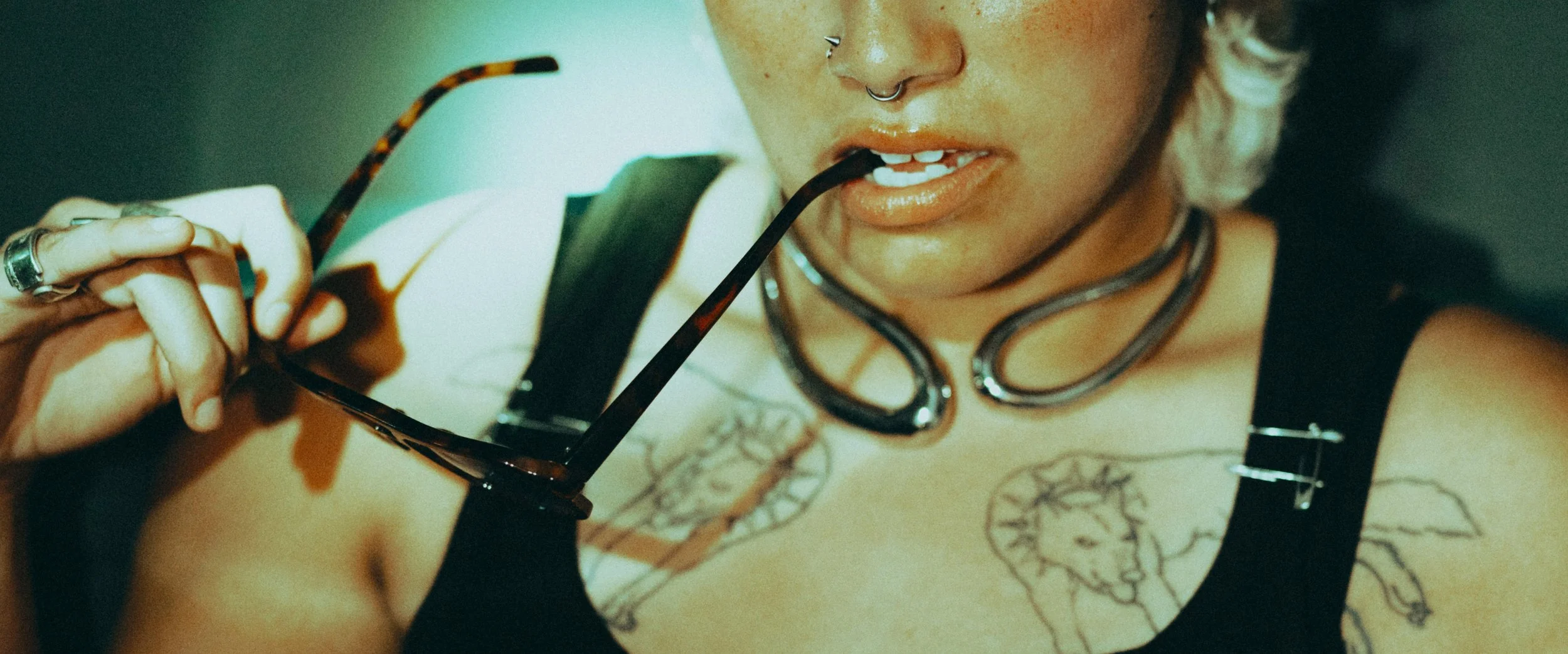 Close-up of a woman with tattoos, nose piercings, and layered necklaces, holding sunglasses with tortoiseshell frames and biting the arm of her sunglasses.
