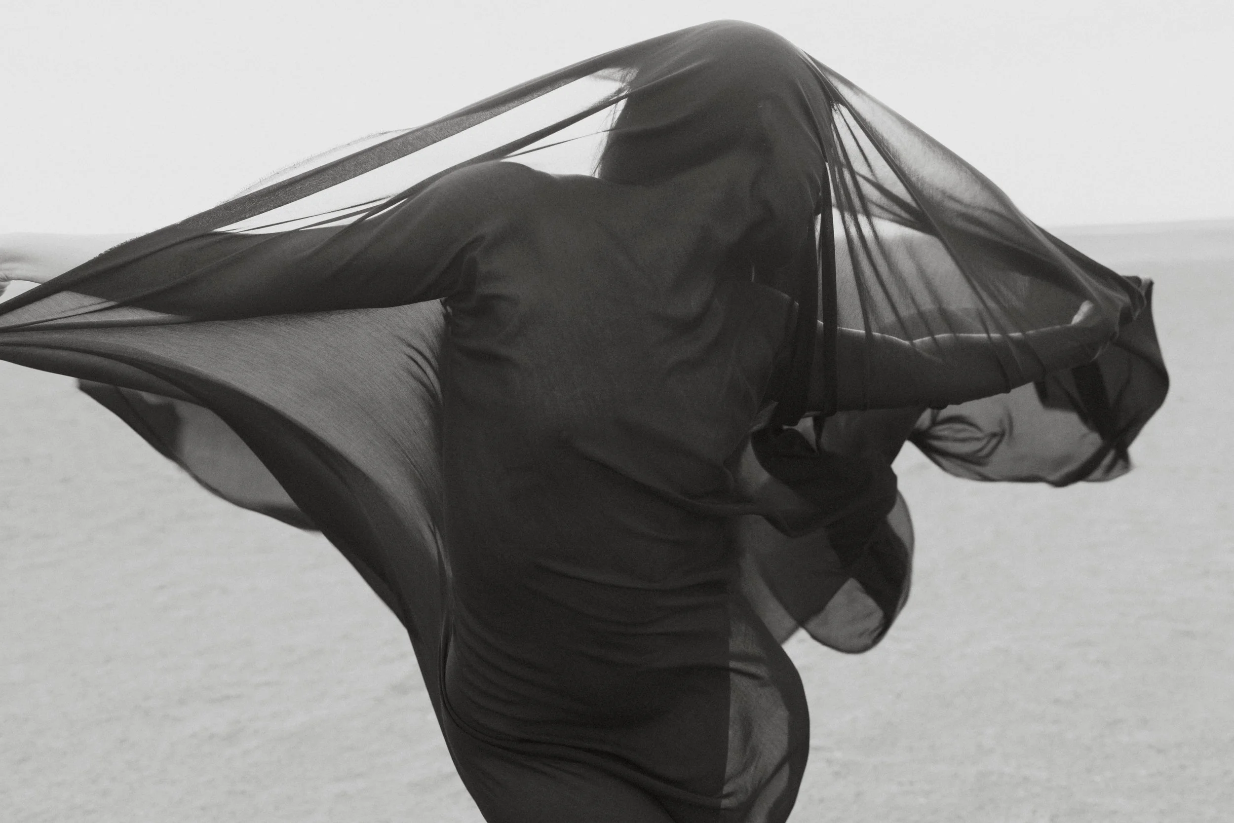 A person wearing a black dress with sheer, flowing fabric, posing on a beach with a gray, overcast sky in the background.