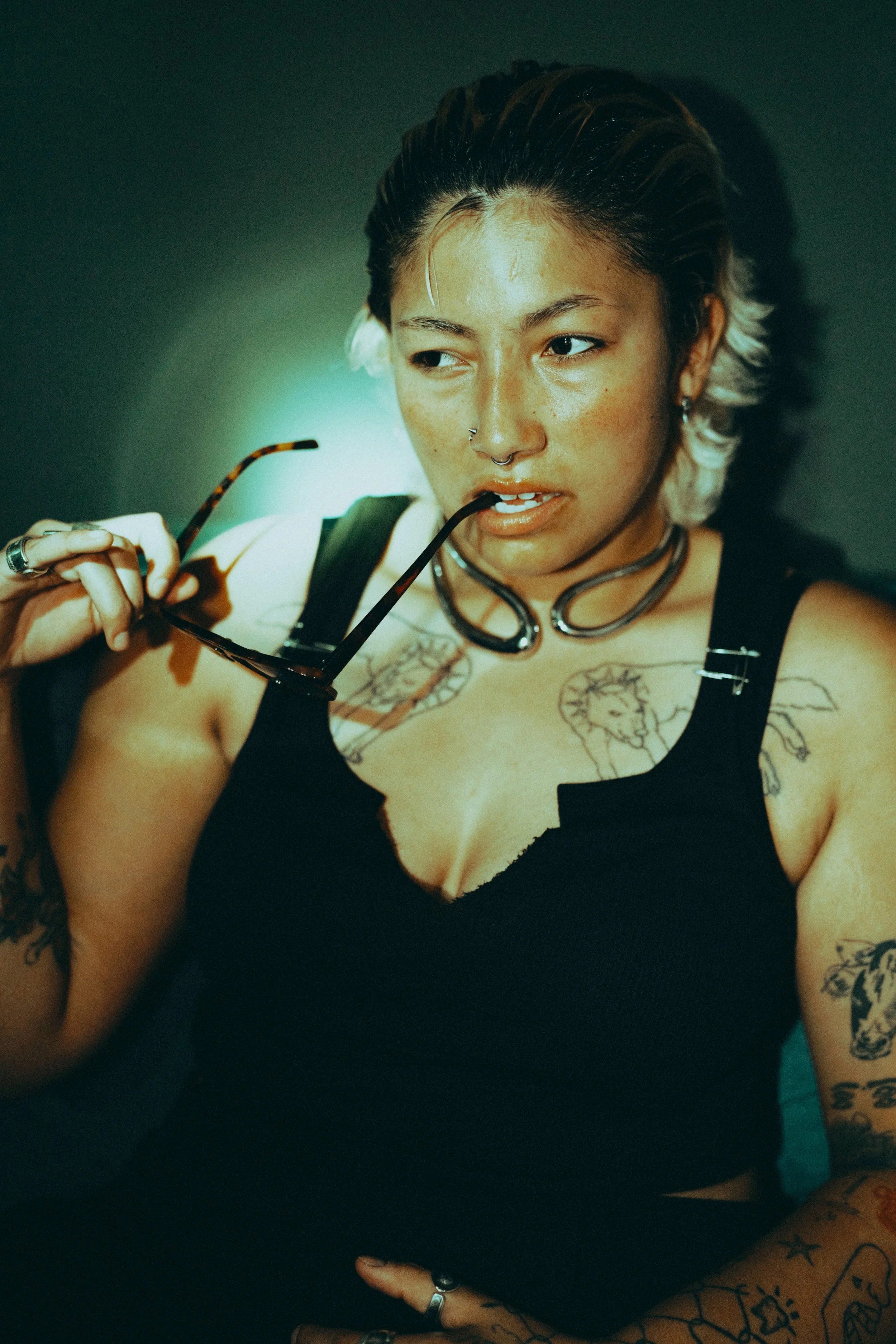 A woman with tattoos on her chest and arms, wearing a black top, holding sunglasses in her hand, and biting the arm of her glasses, with dark hair and multiple piercings.