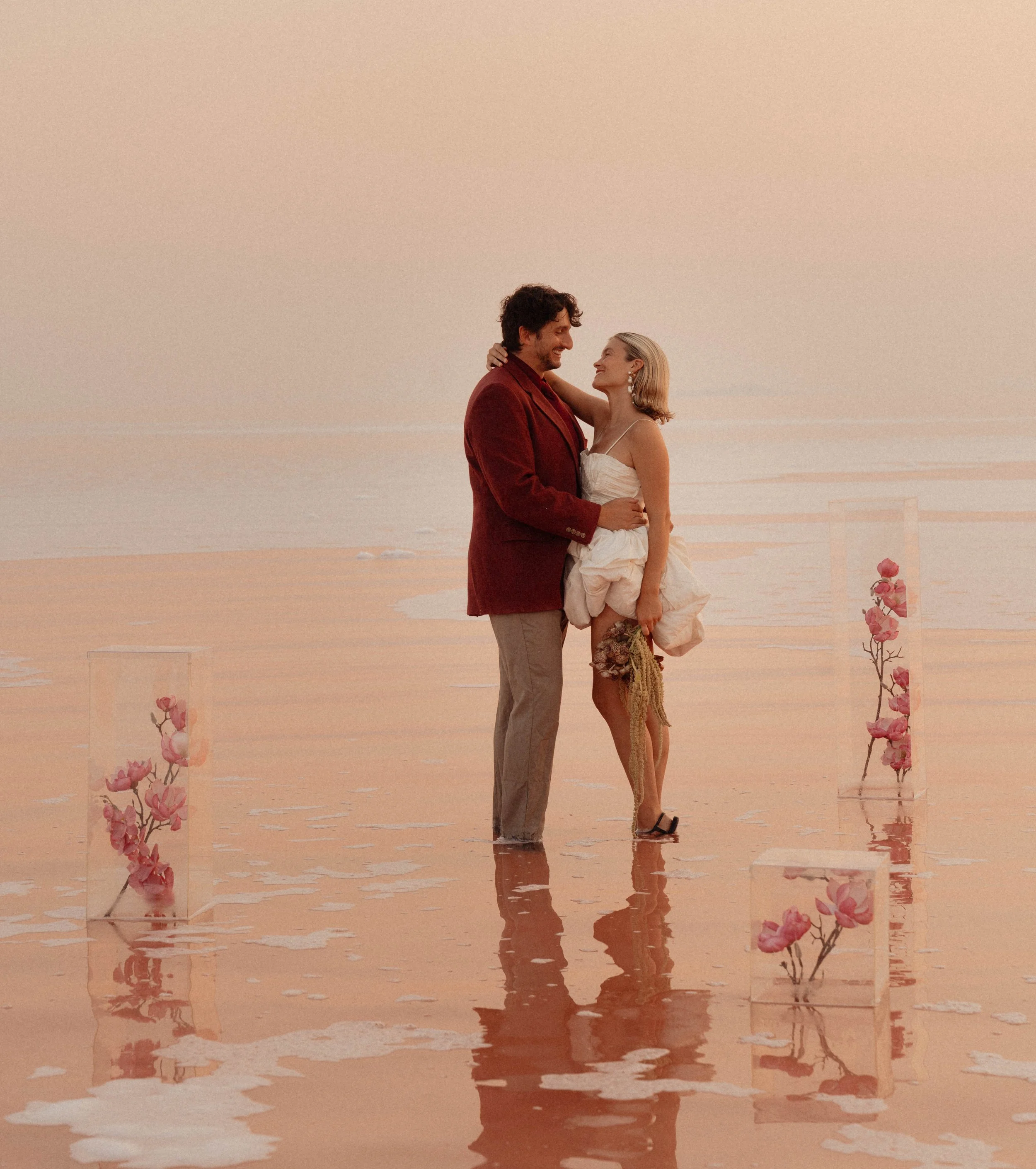 A couple stands in shallow water at the beach during sunset, embracing and gazing into each other's eyes. The woman wears a white dress and earrings, and the man wears a red blazer and light trousers. There are pink flowers in tall vases on either si