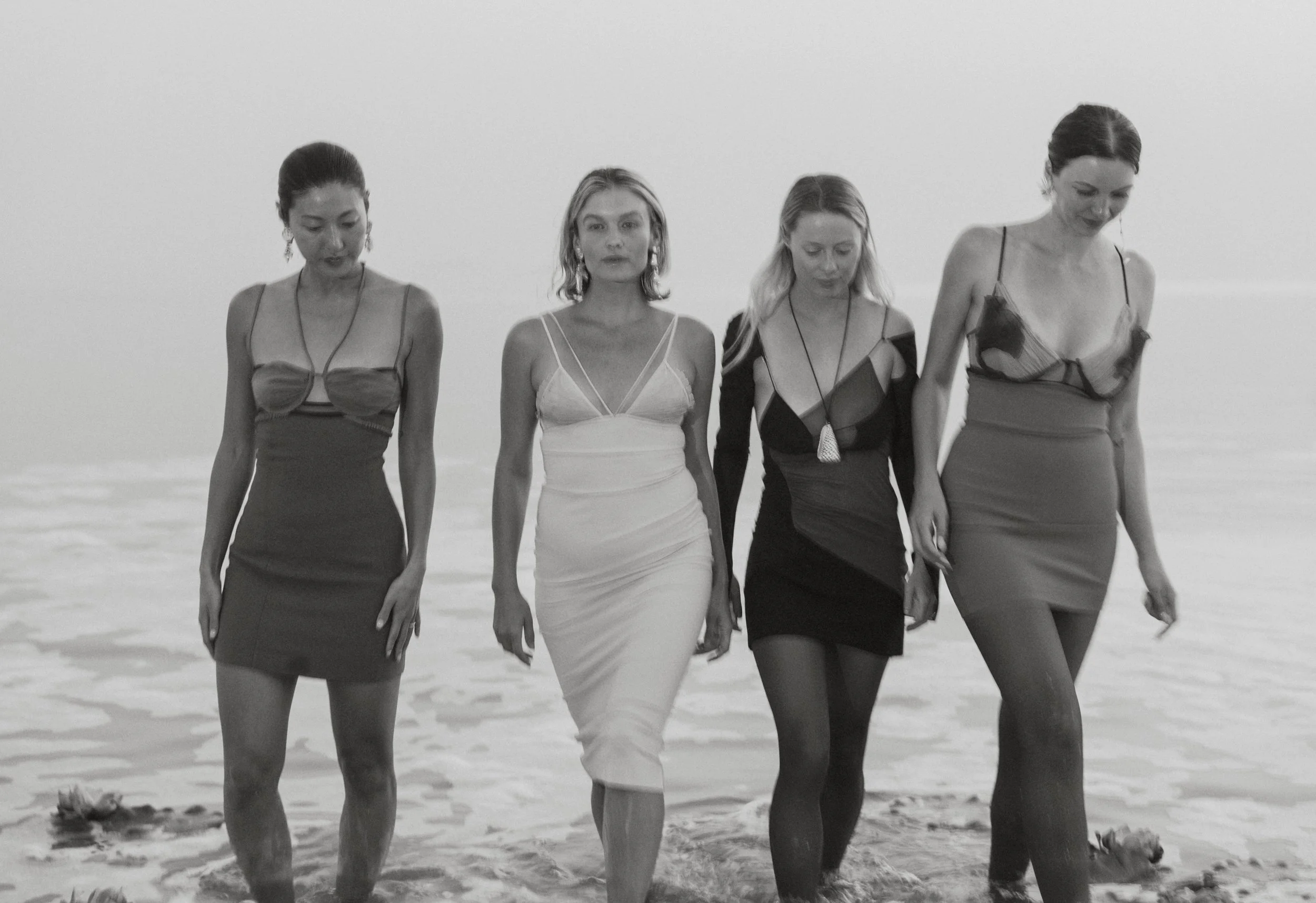 Four women walking on a beach in a line, dressed in elegant dresses, with the ocean in the background in black and white.