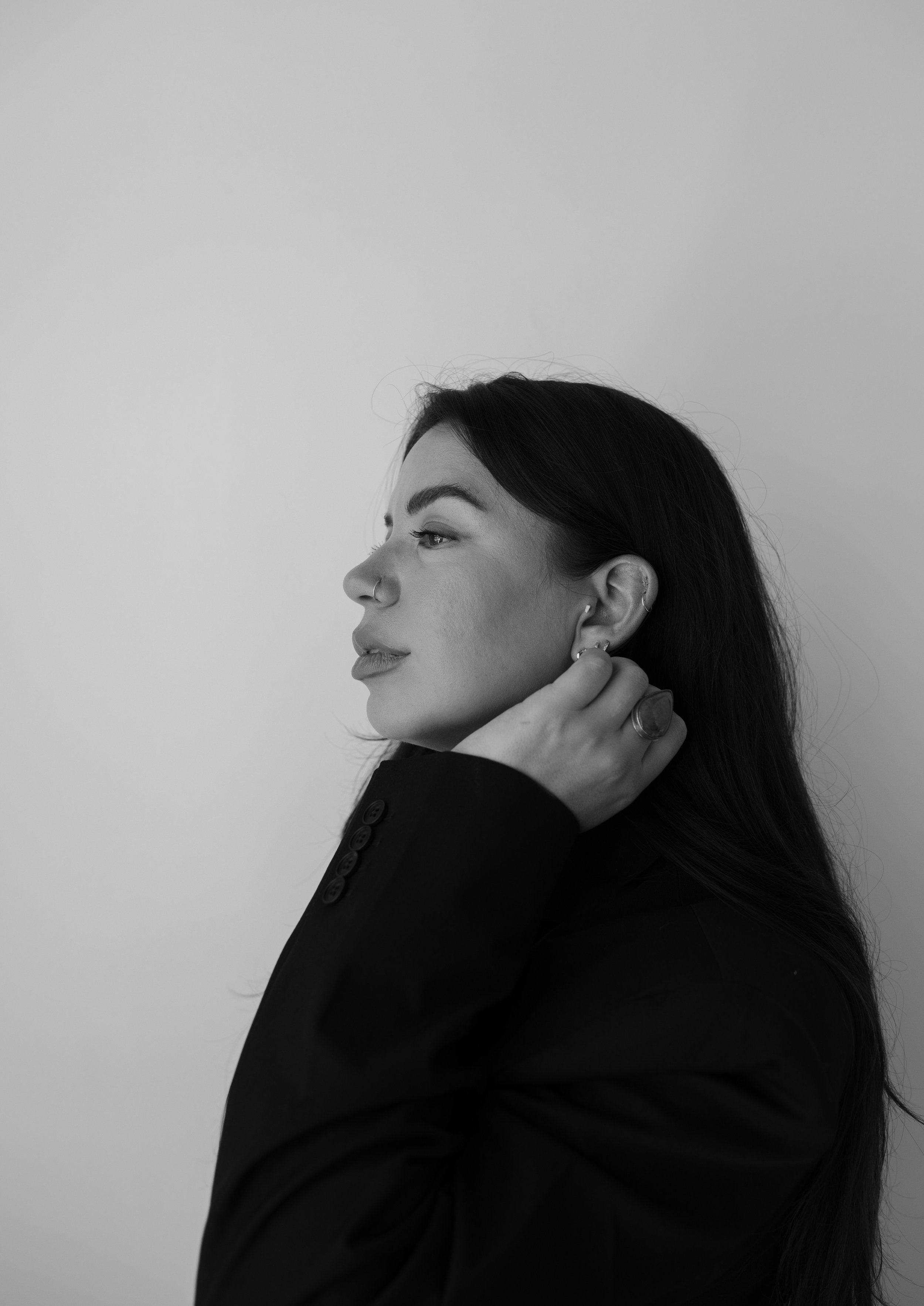 Black and white side profile of a woman with dark hair, wearing rings and earrings, touching her neck, against a plain light background.
