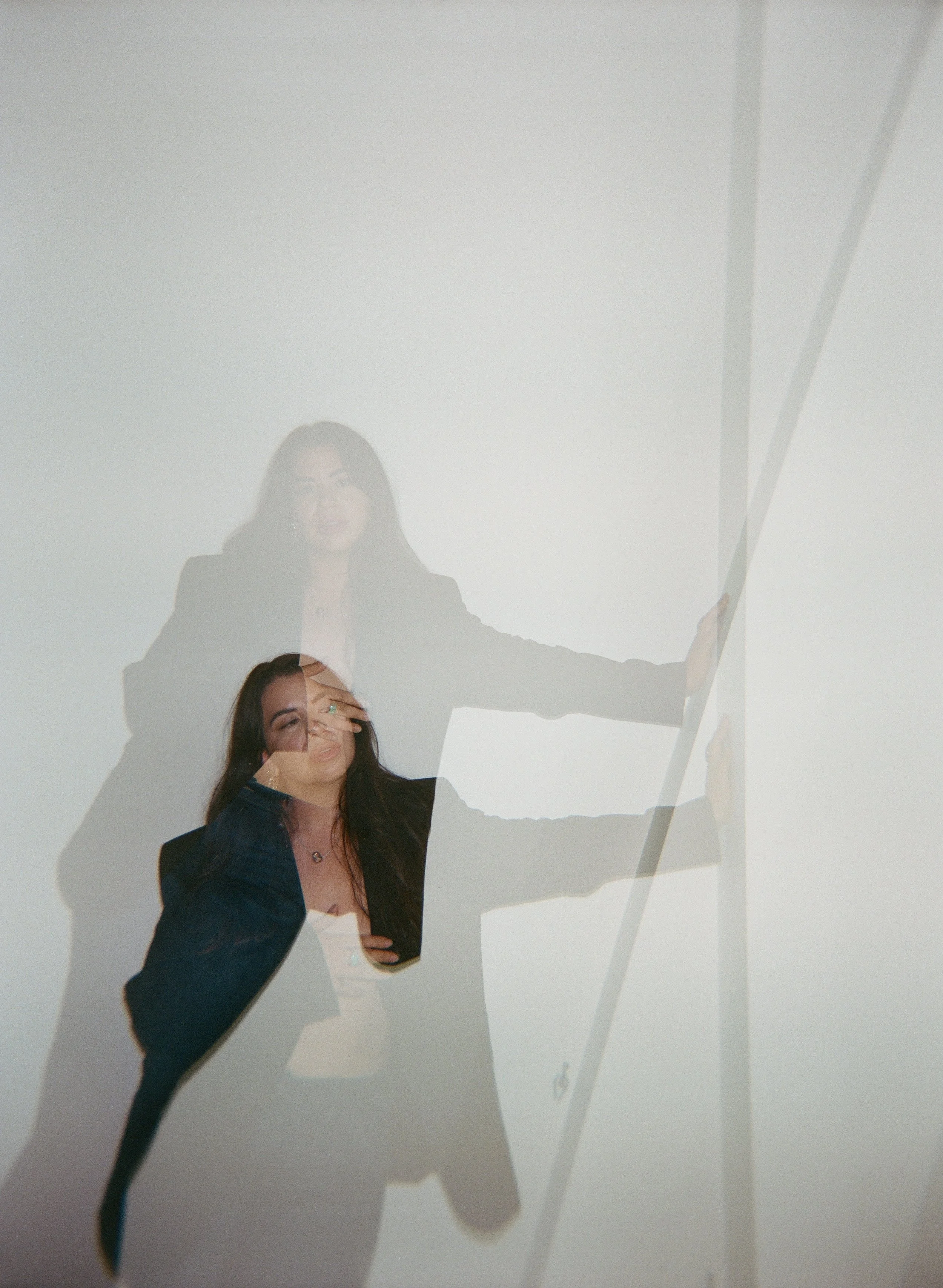 The image shows two women, one with dark hair in a black blazer and the other with lighter hair, wearing a dark jacket, posed against a light-colored background. Their shadows and reflections are visible on the surface behind them.