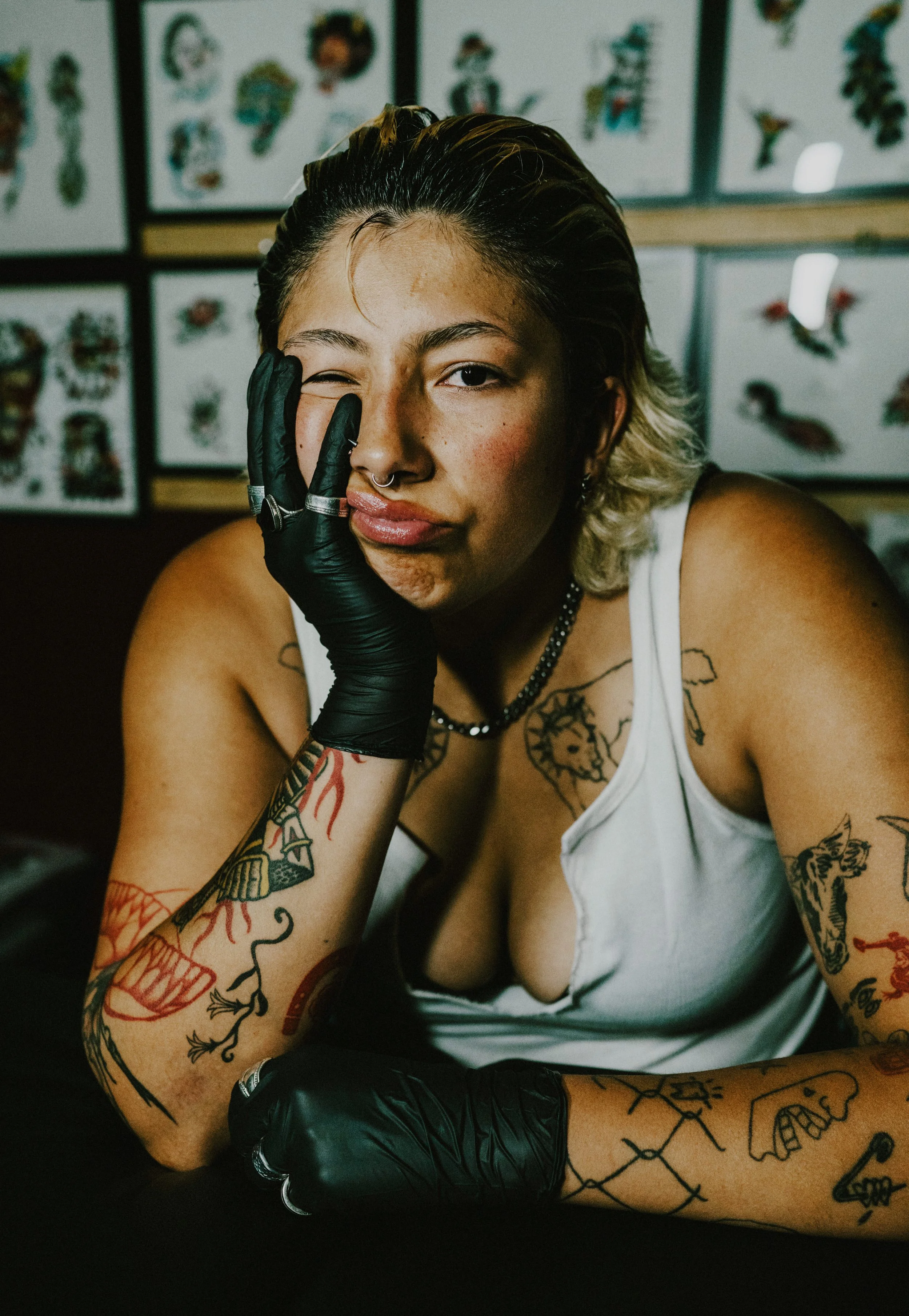 A woman with tattoos, wearing a white tank top, black gloves, and jewelry, winking with her hand on her face, in front of tattoo flash art on the wall.