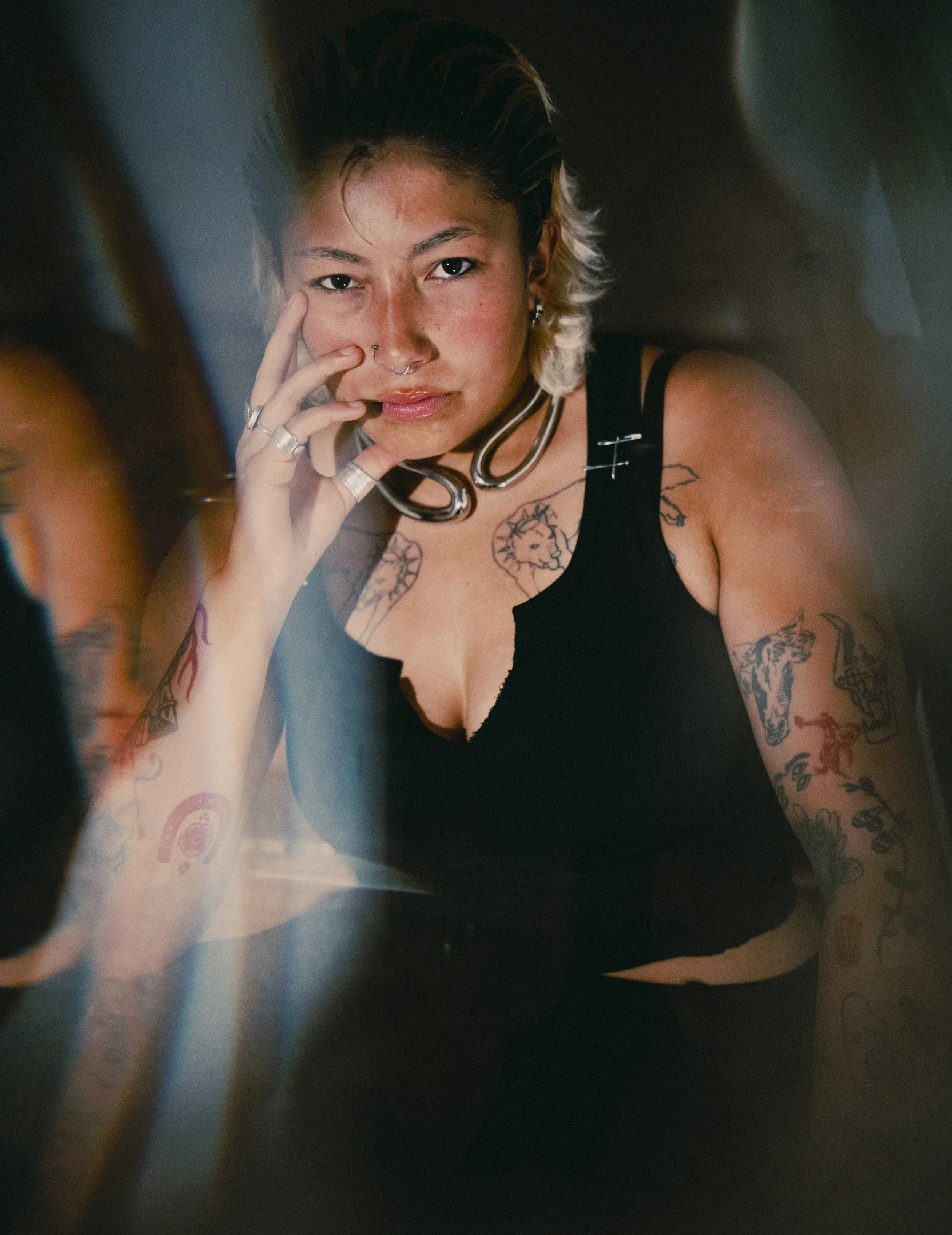 A woman with short, dark blonde hair with lighter tips, tattoos on her arms and chest, wearing a black top, jewelry including rings, earrings, and a choker, and a septum nose ring, looking into a mirror with her hand near her face.