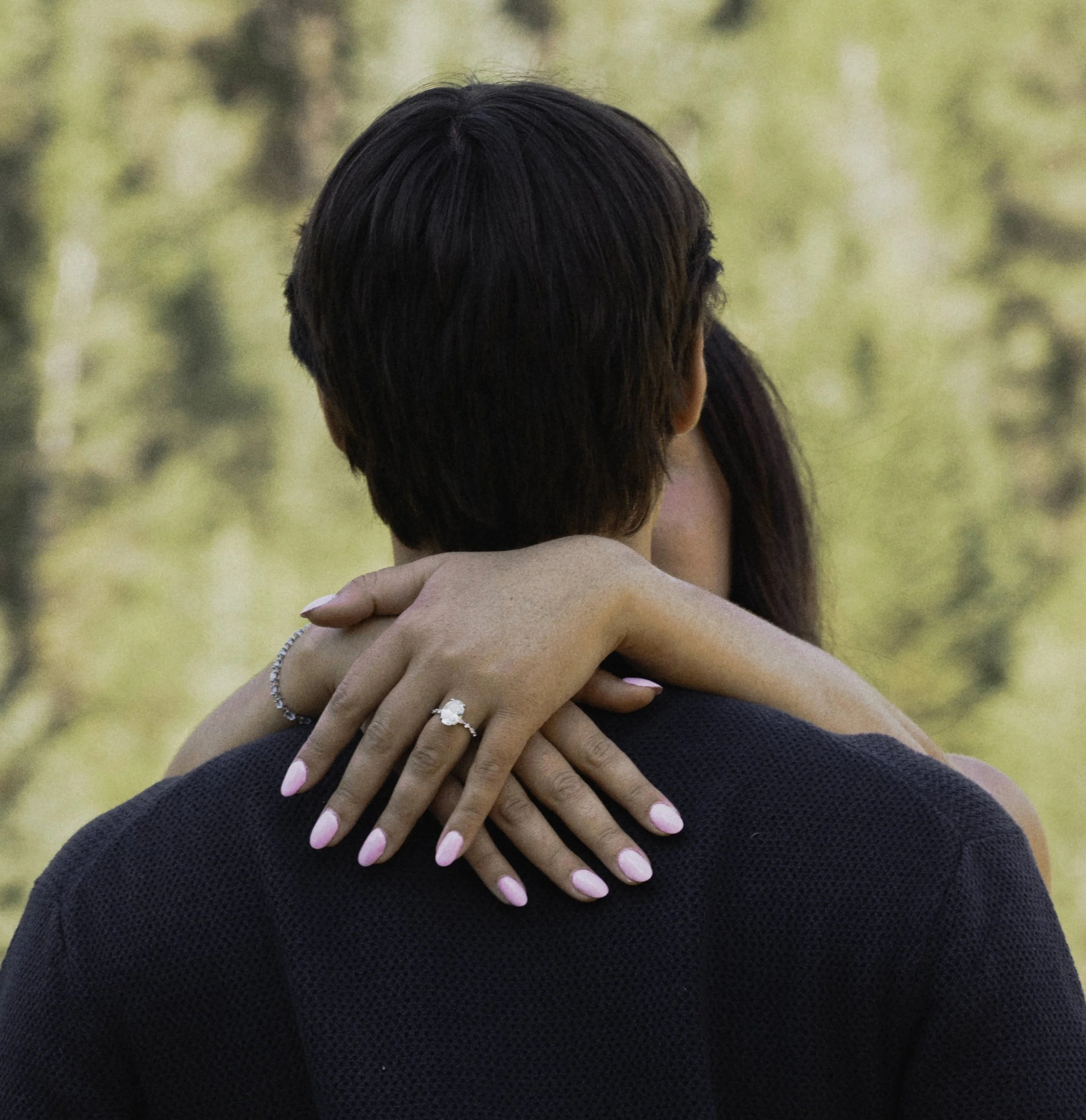 Back of woman's head with short, dark hair and her hand with pink manicured nails resting on another person's shoulder, outside in a blurred natural background.