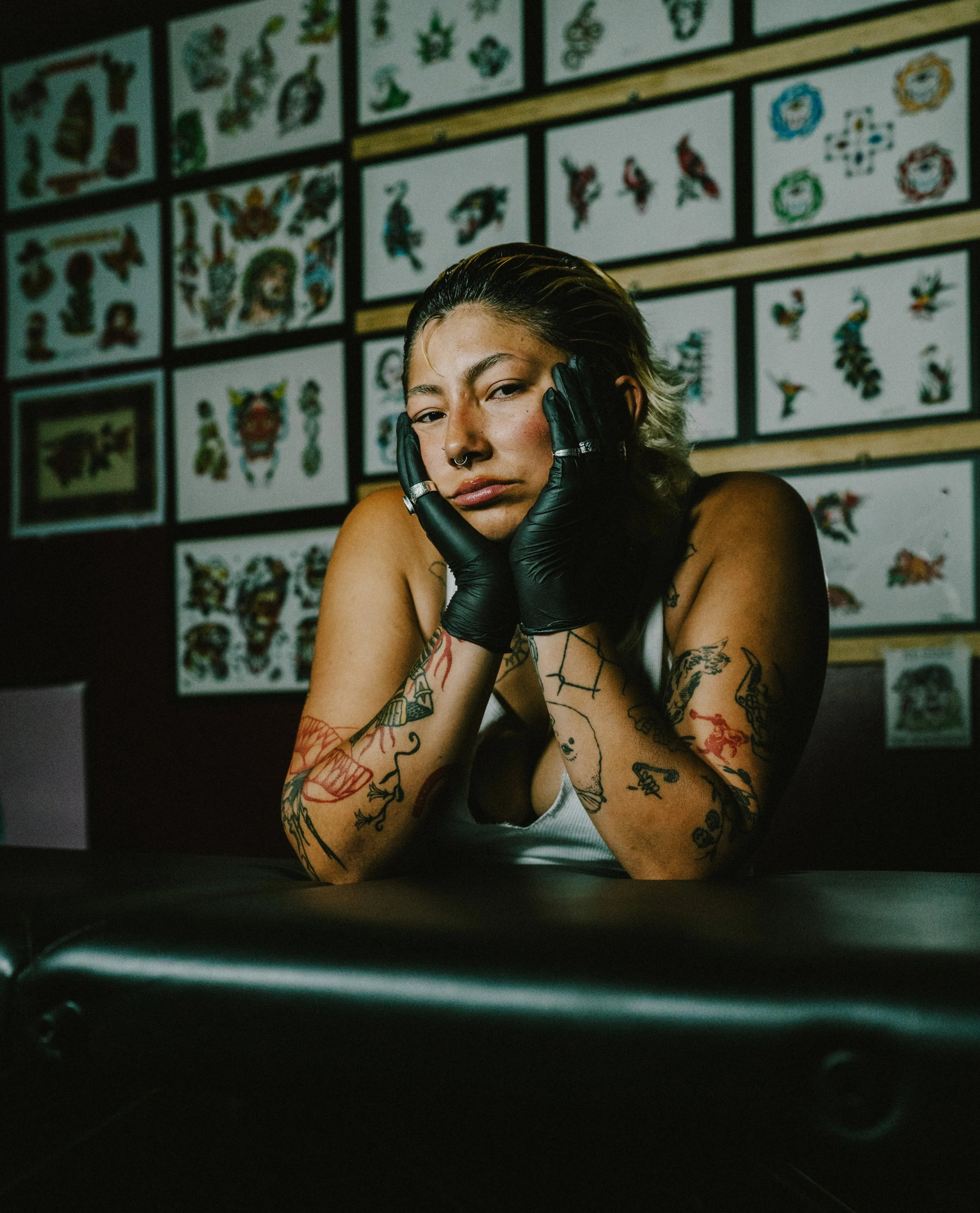 A woman with short, dark hair and tattoos on her arms, wearing black gloves, resting her face in her hands, sitting in front of a wall decorated with numerous tattoo designs.