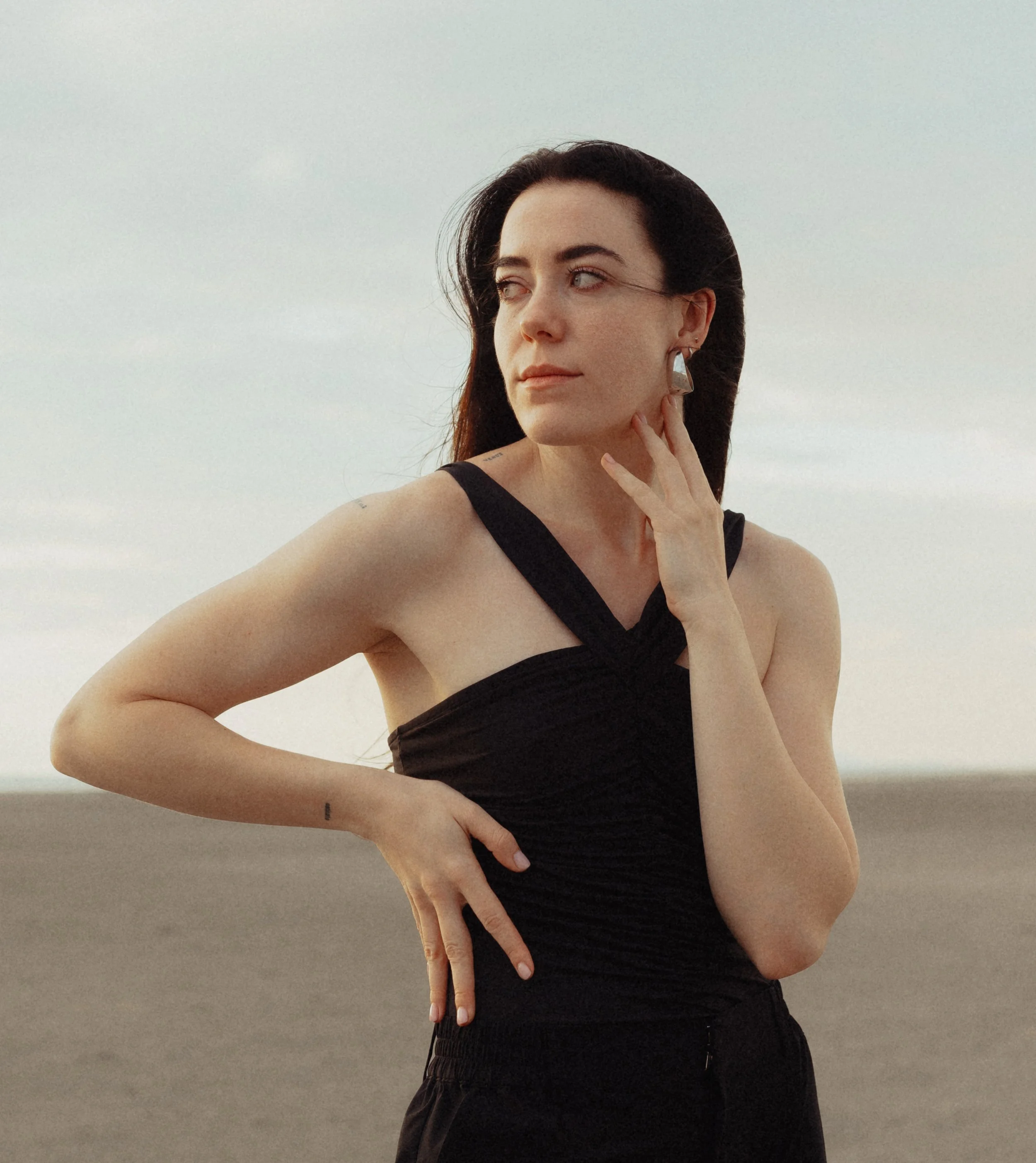 A woman with long dark hair, wearing a black sleeveless top and large earrings, touching her face with her right hand, standing outdoors against a cloudy sky.