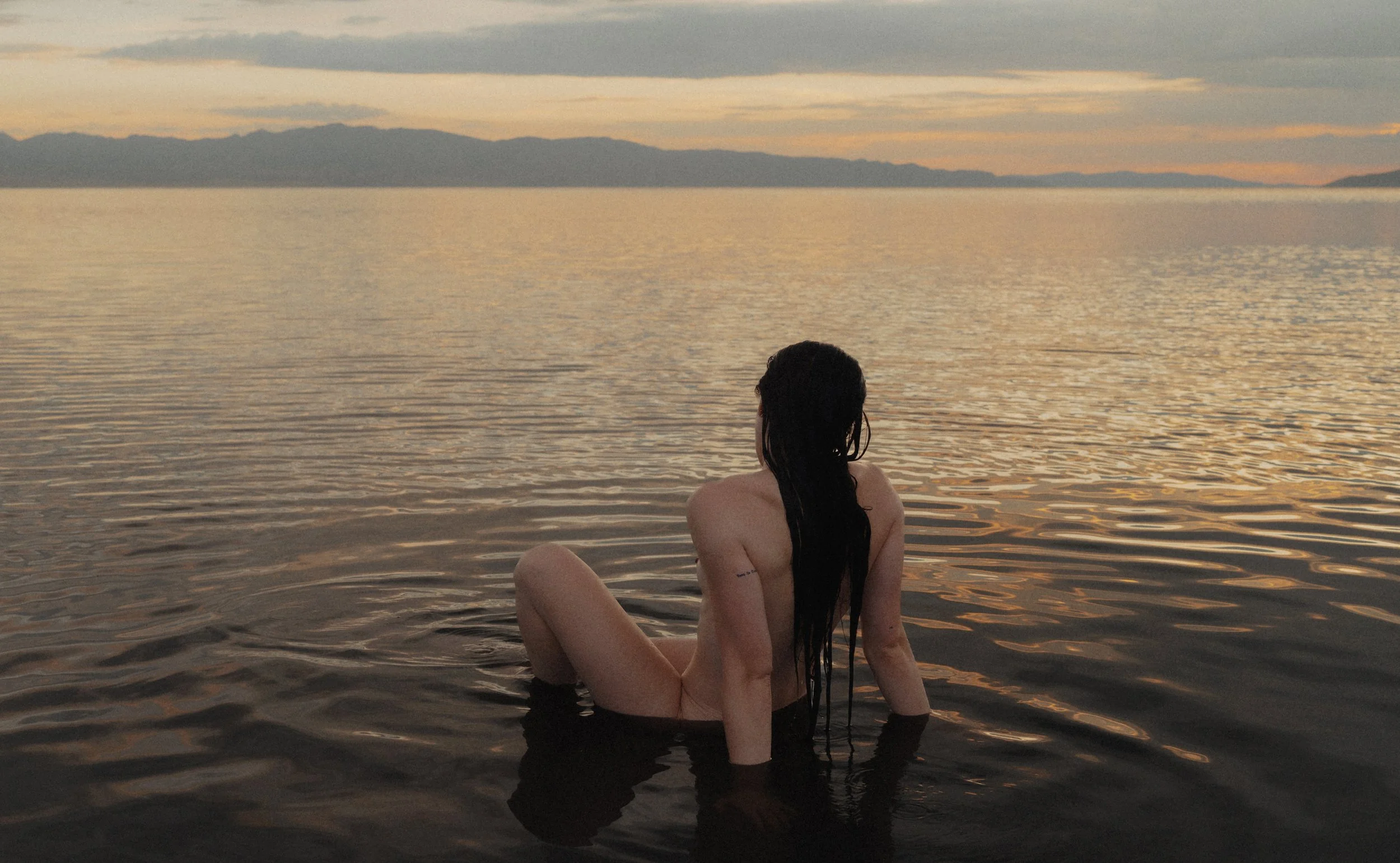 A woman with long dark hair in a bath, partially submerged, sitting in body of water at sunset or sunrise, with mountains and cloudy sky in the background.