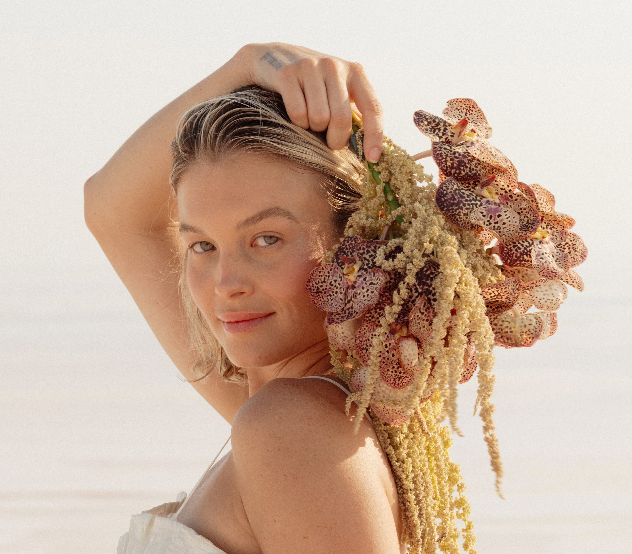 A person with blonde hair and hazel eyes posing with their hand on their head, wearing a white top, and surrounded by large yellow and purple orchids against a plain light background.