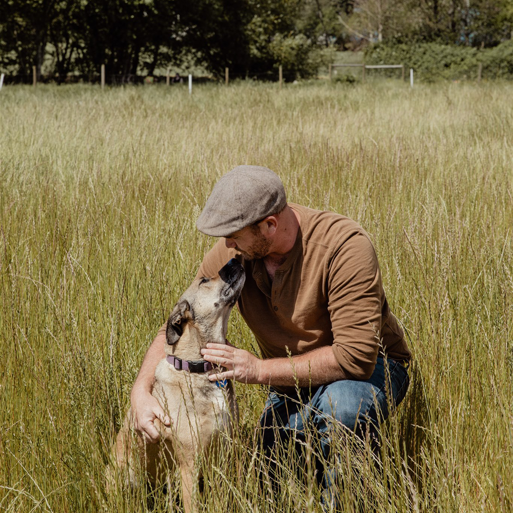 A man kneeling in a grassy field, hugging and nuzzling a dog.