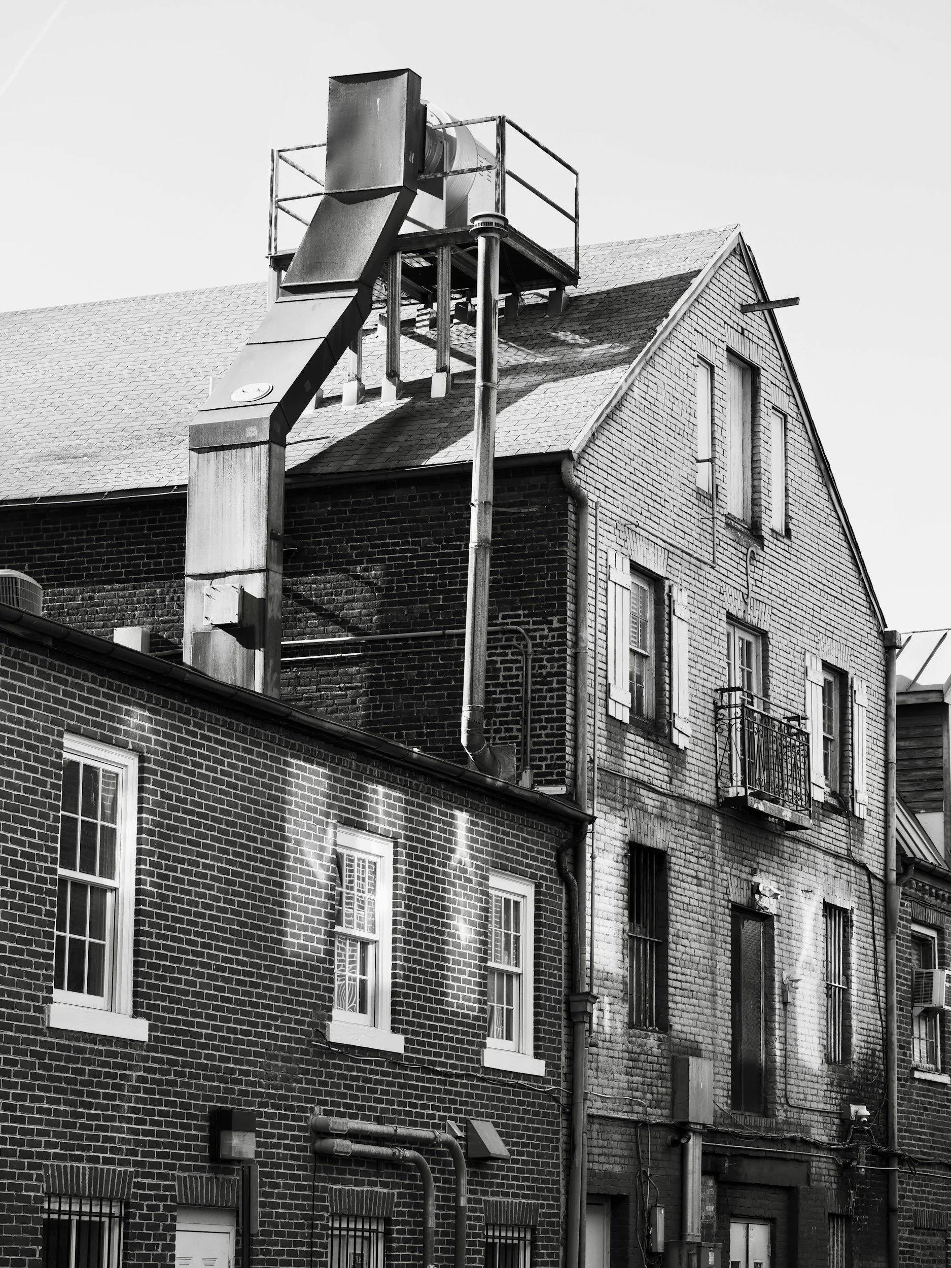 A black and white photo of a multi-story brick building with several windows, some with metal bars, and a prominent chimney pipe extending from the roof with a vent at the top. The building has a sloped roof and visible pipes and wires along the exterior walls.
